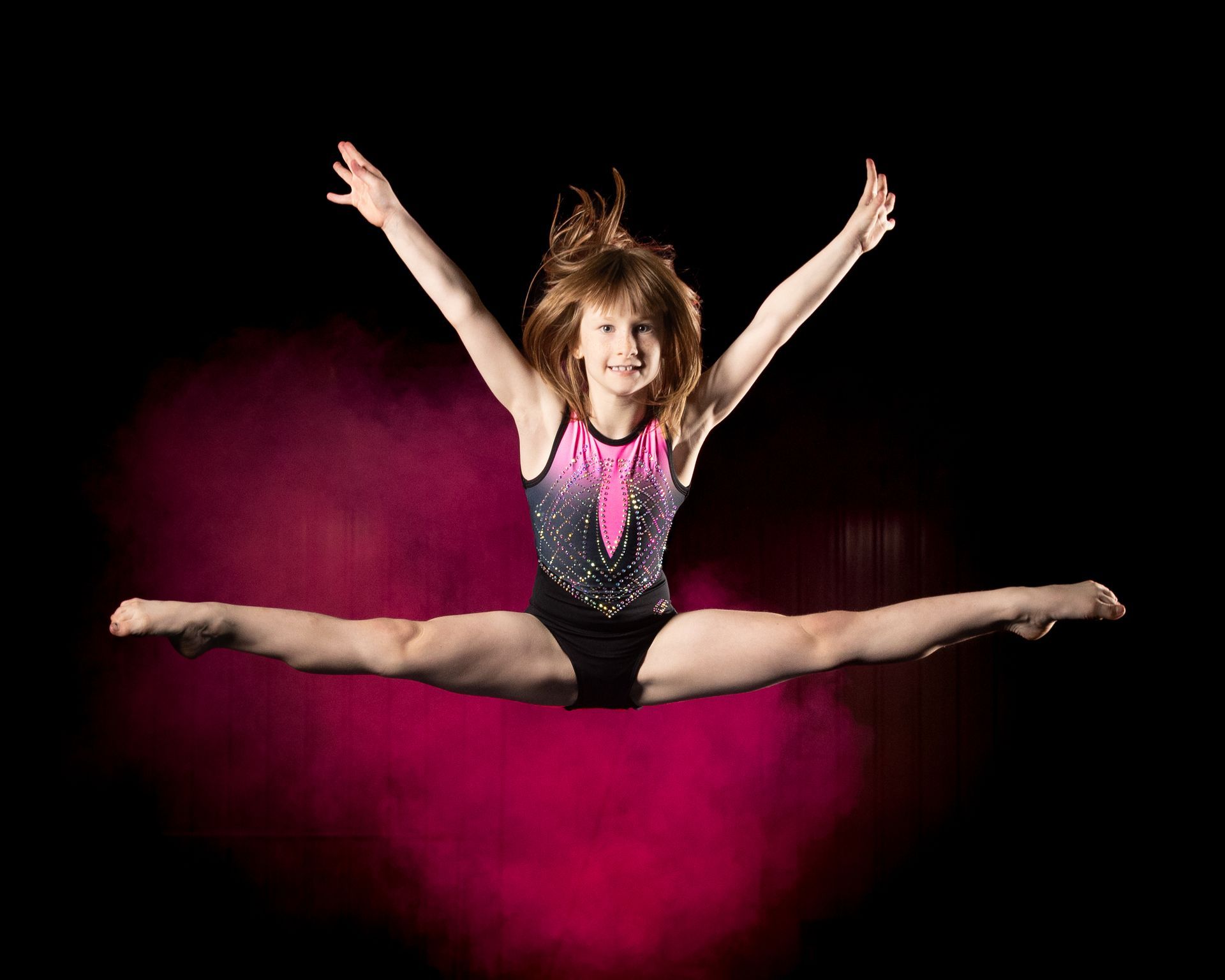 Gymnast in a straddle jump, arms outstretched, smiling against a dark background with pink haze.
