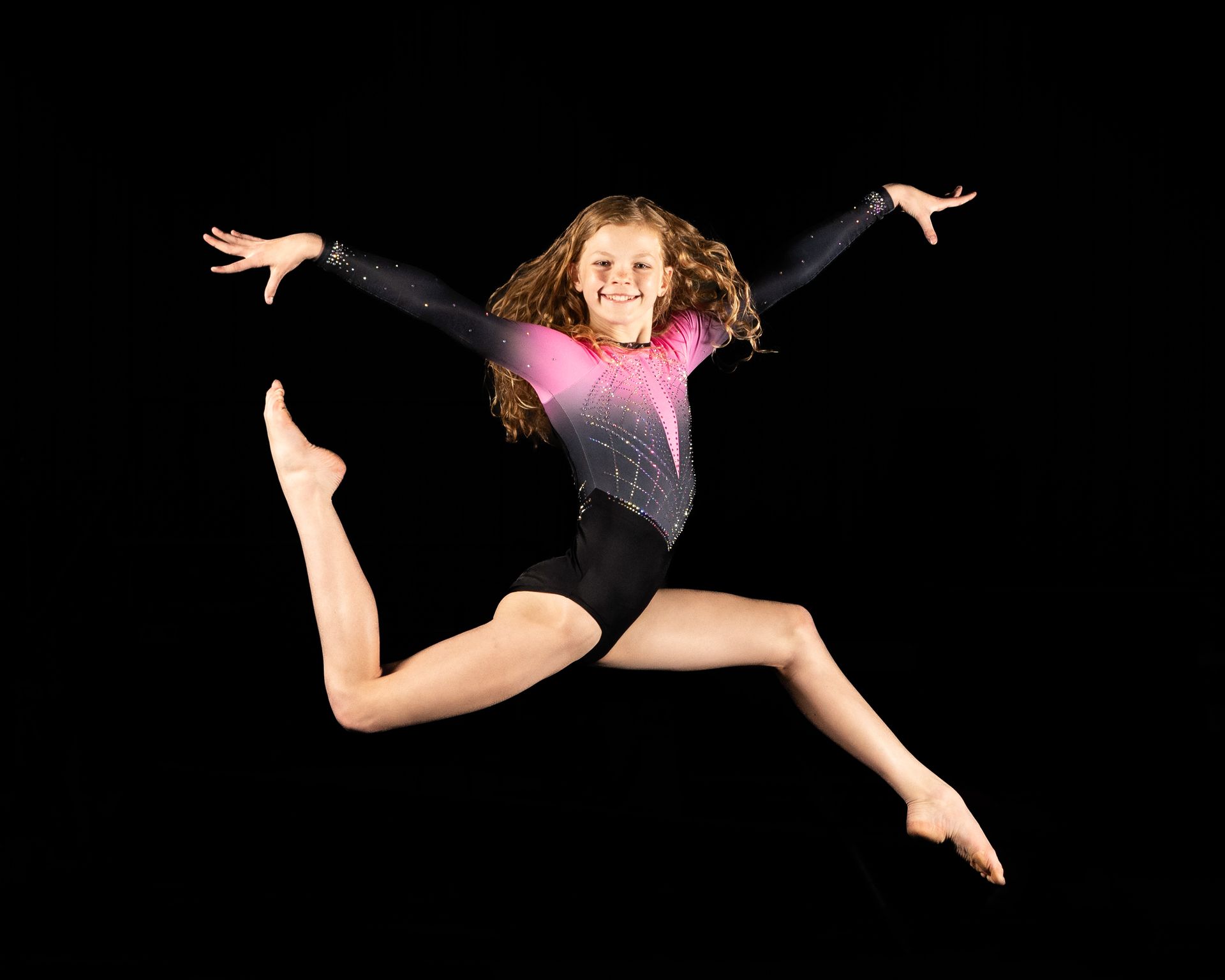 Gymnast in a black and pink leotard mid-air, arms and legs spread, smiling against a black background.