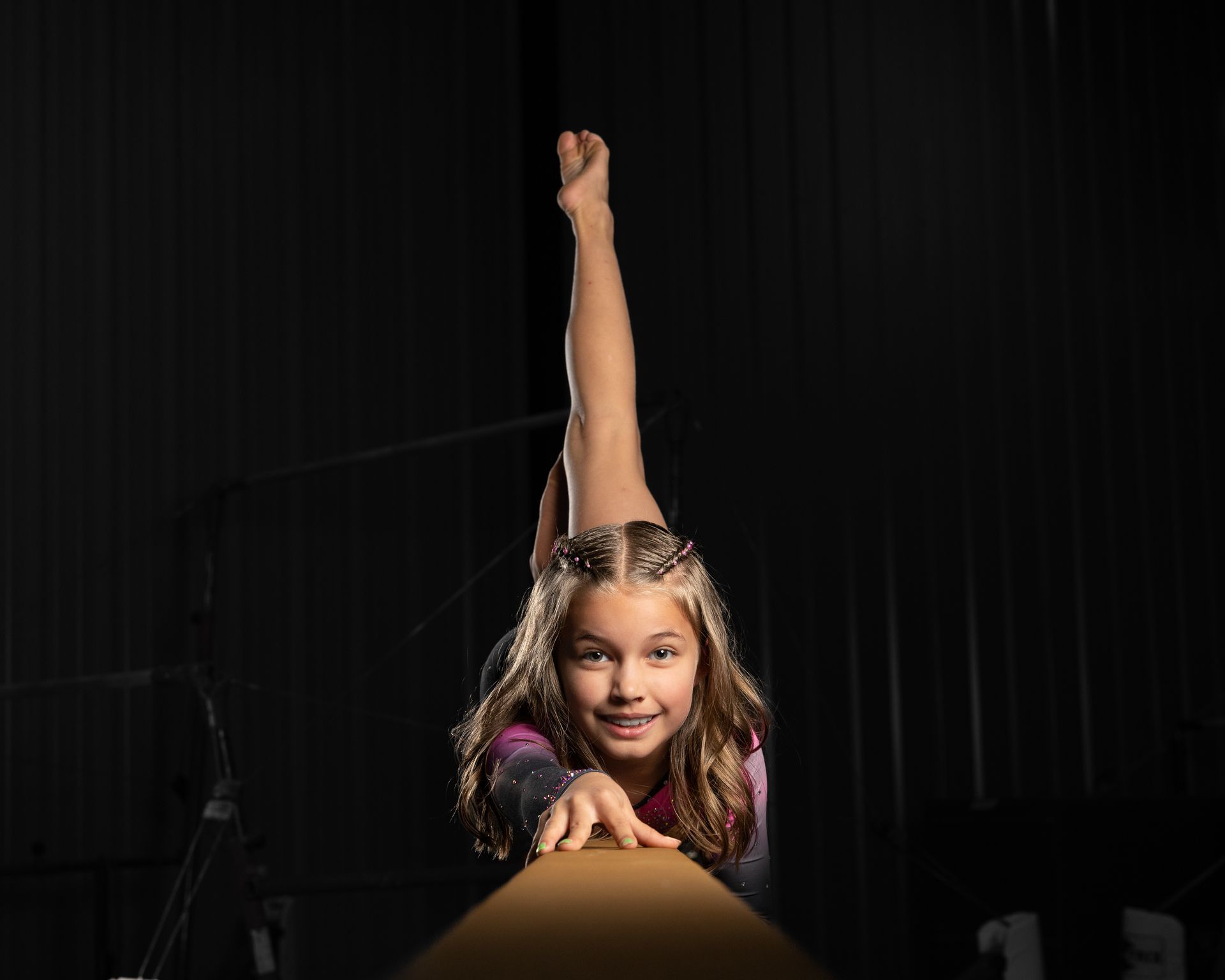 Gymnast balances on a beam, leg extended. She smiles, wearing a leotard against a dark backdrop.