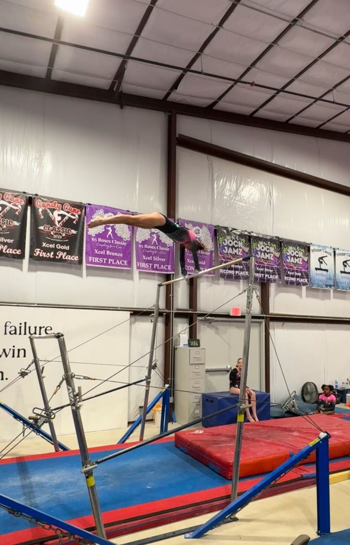 Gymnast on uneven bars, mid-flight, arms extended, legs straight. Indoor gym setting with purple banners.