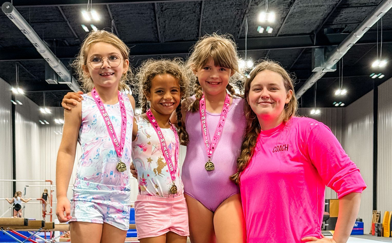 Four young gymnasts with medals pose with a coach, smiling in a gym.