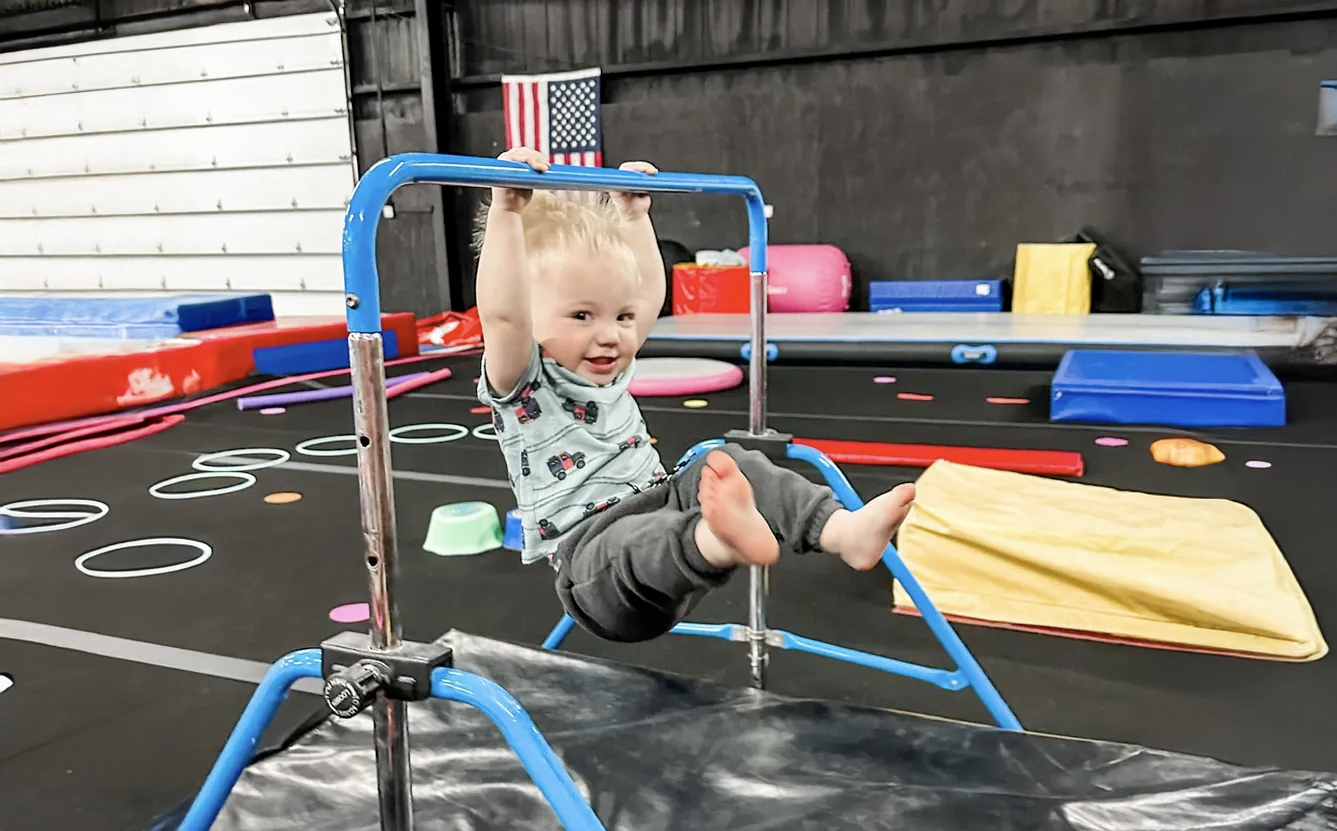 Child hanging from a blue gymnastic bar, smiling, in a gym setting with mats and equipment.