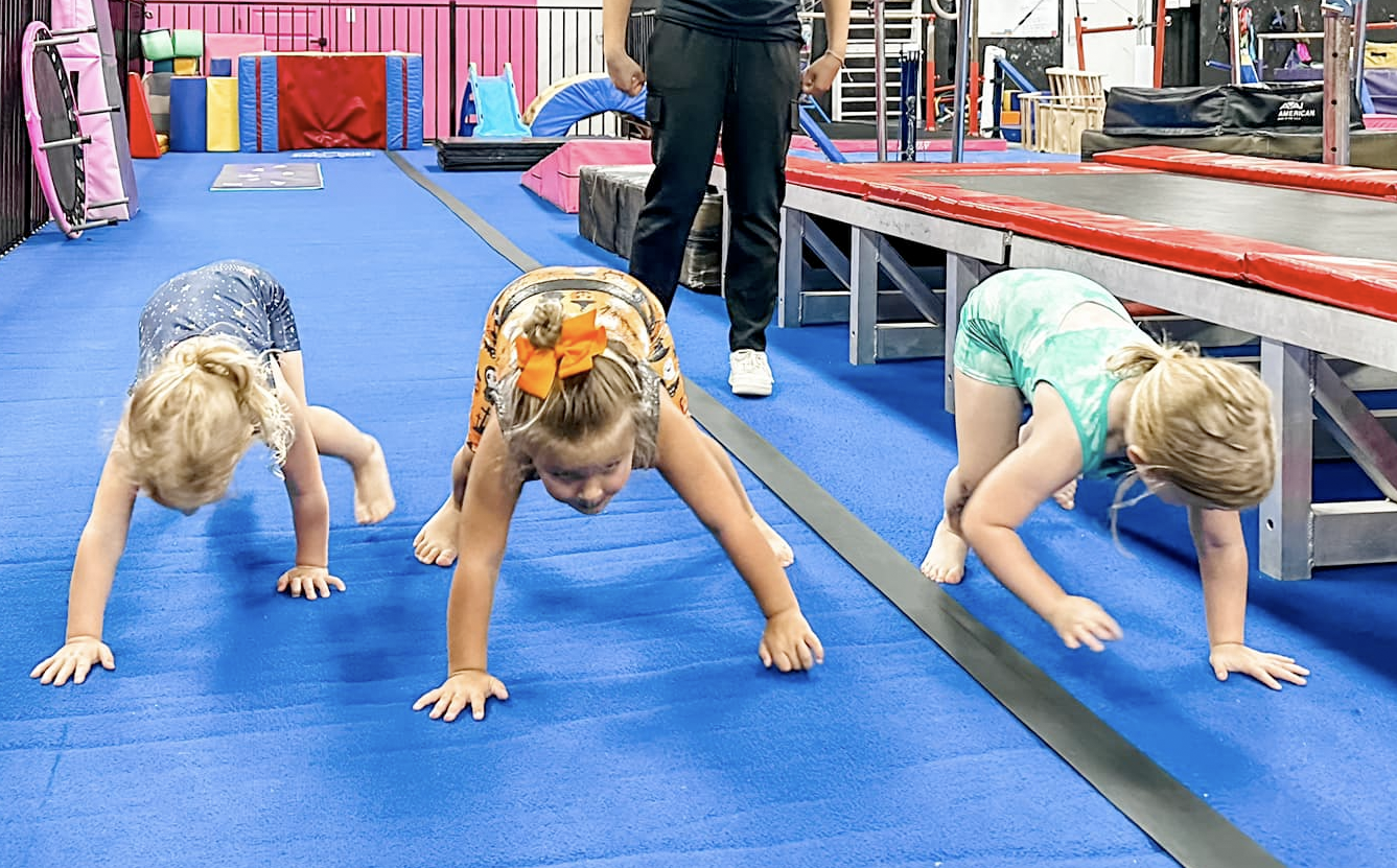 Three young children crawling on a blue mat in a gymnastics gym, supervised by an adult.