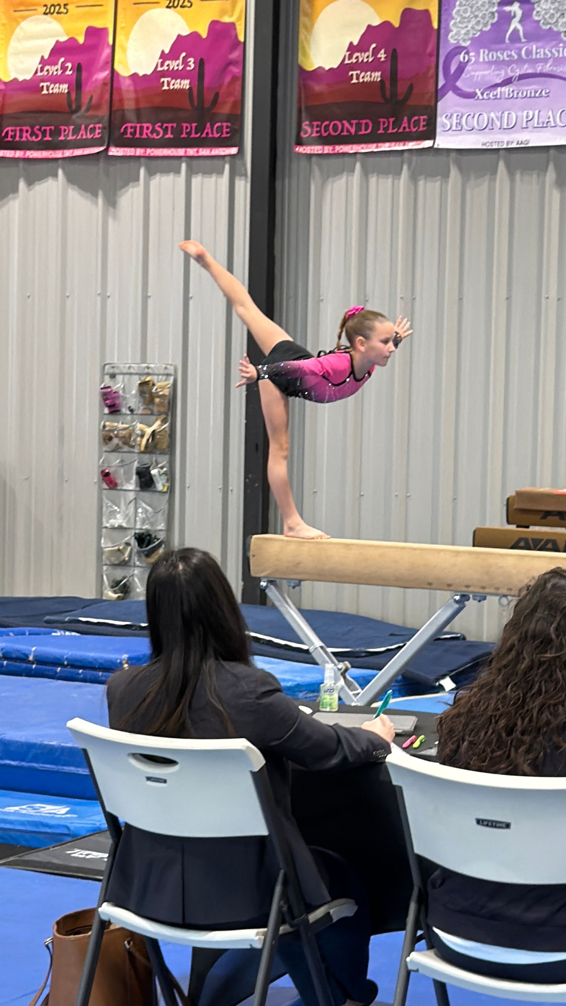 Gymnast in pink leotard on balance beam, arm and leg extended, in a gymnasium.