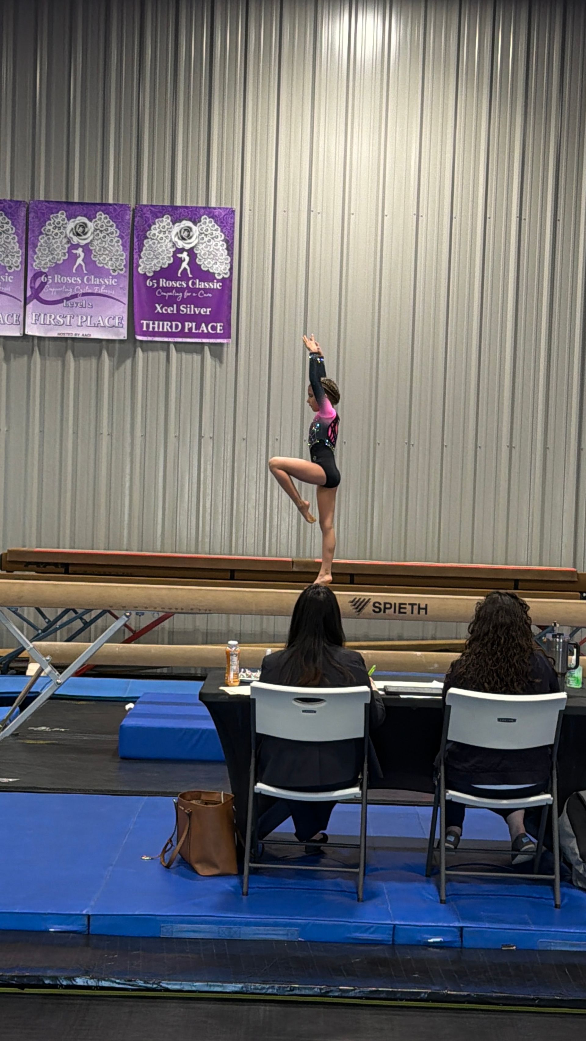 Gymnast balances on a beam, arms raised, in a competition setting.
