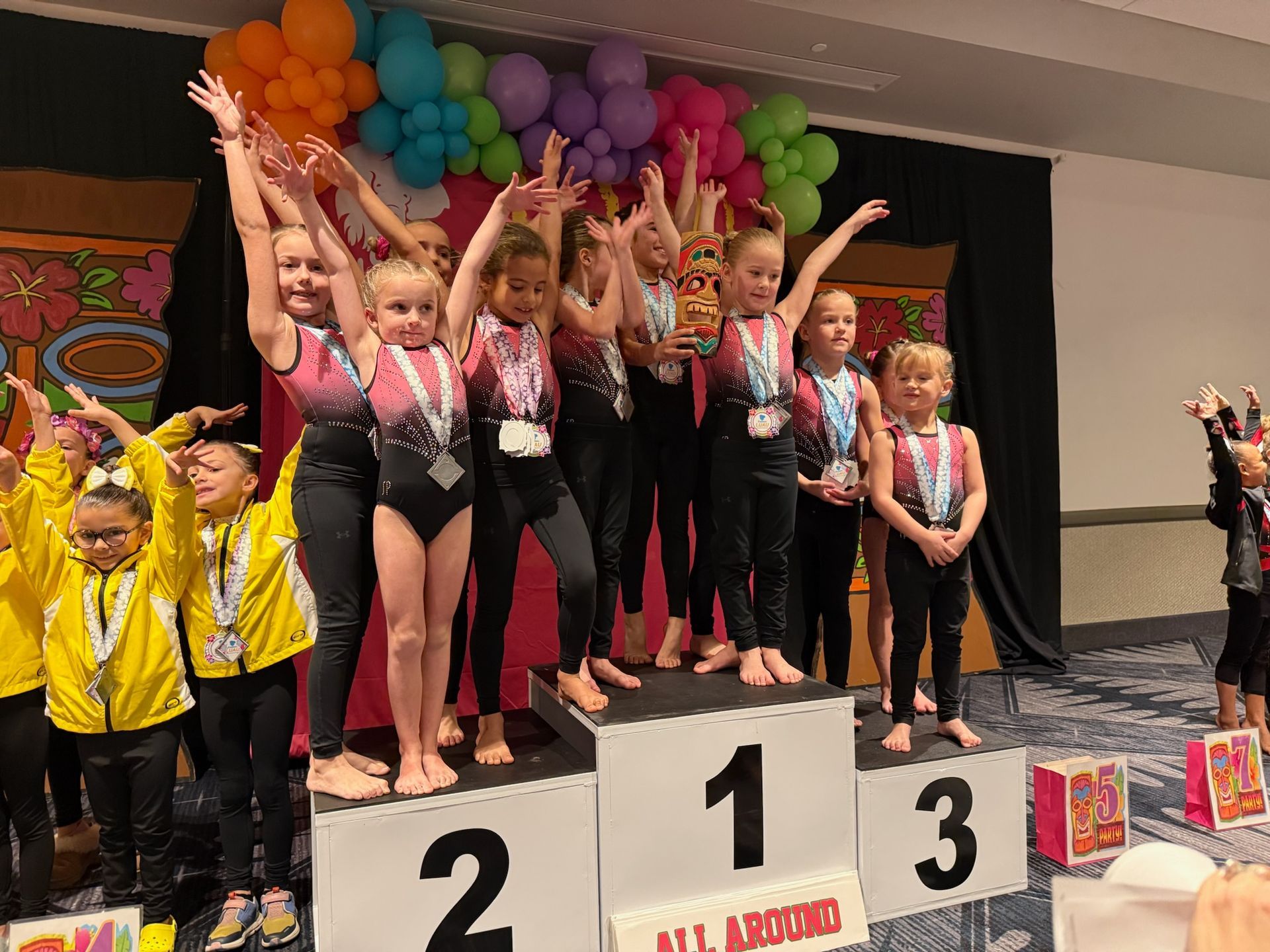 Gymnasts on a podium, wearing leotards and medals, celebrating a win with arms raised.