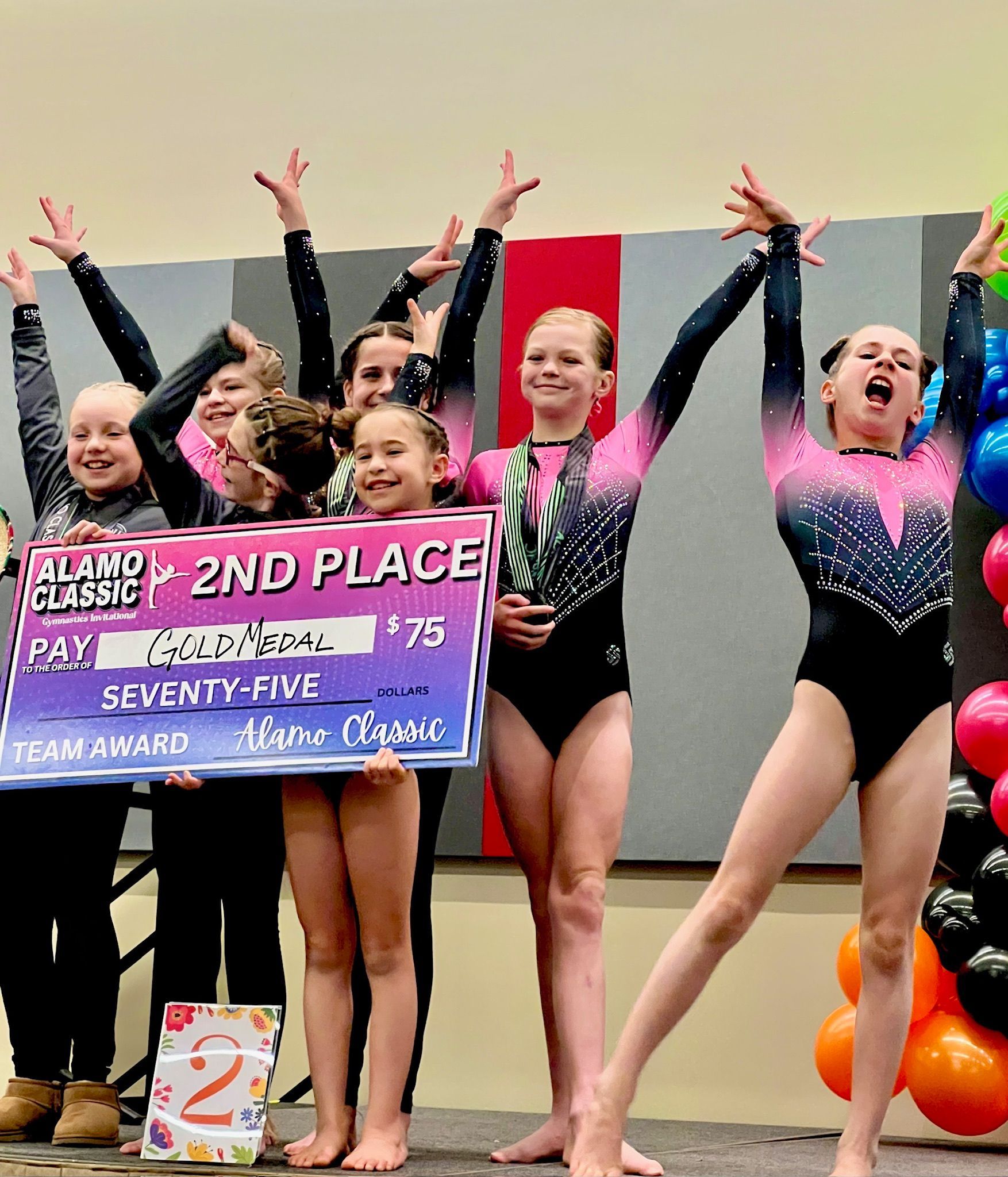 Gymnastics team celebrating 2nd place win at a competition; holding a large check, arms raised, smiling.