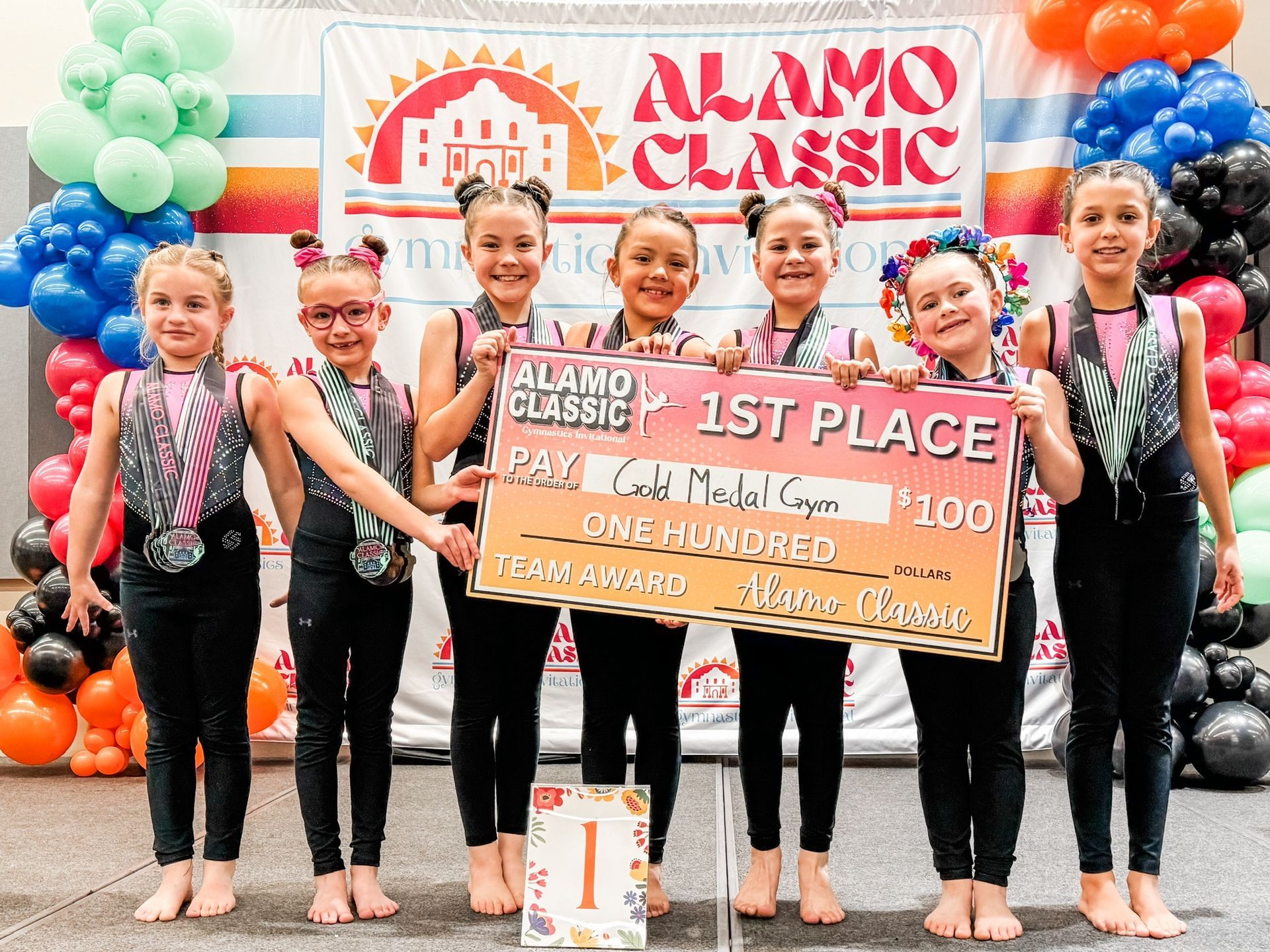 Gymnastics team holding a 1st place check at the Alamo Classic; balloons and event banner in the background.