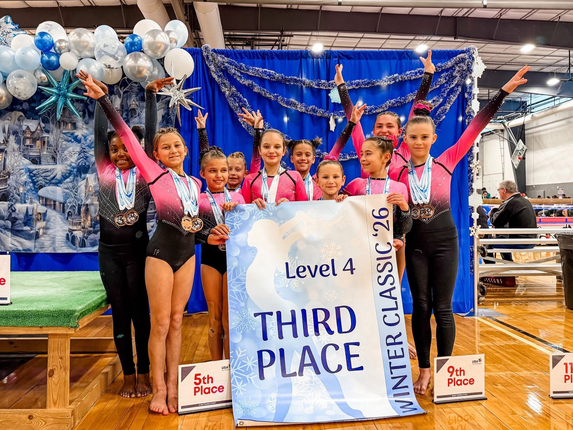 Gymnastics team holding a banner, celebrating a third-place win at Winter Classic '26. They wear pink and black leotards.