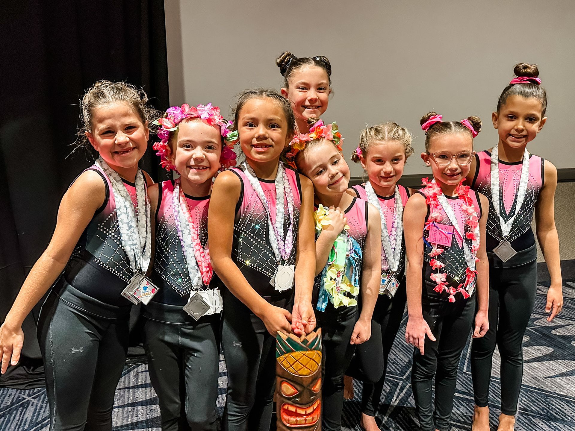 Group of young dancers in black and pink costumes with leis and medals, smiling at a competition.