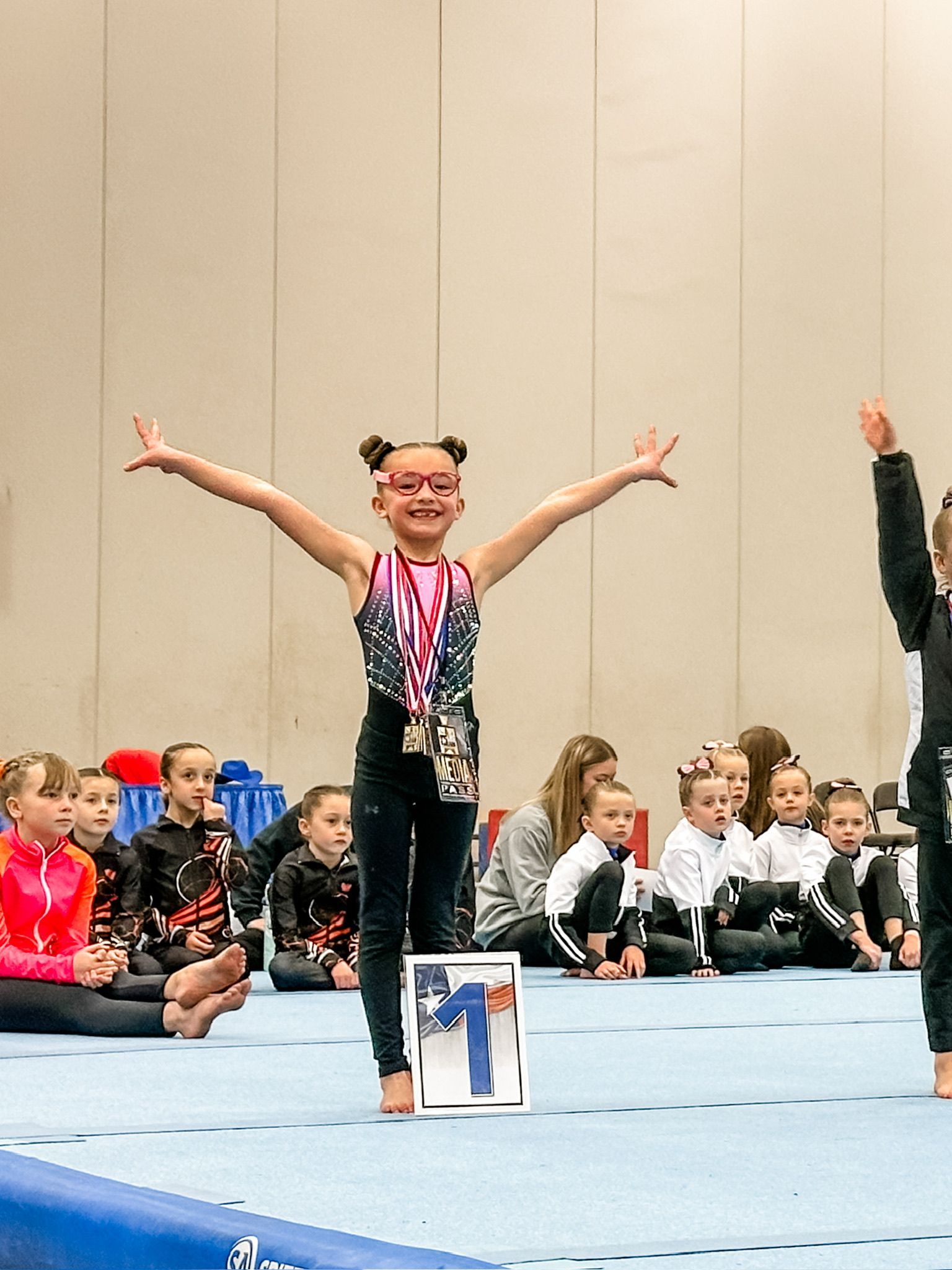 Gymnast with medals and outstretched arms stands on a mat, celebrating. Audience in the background.