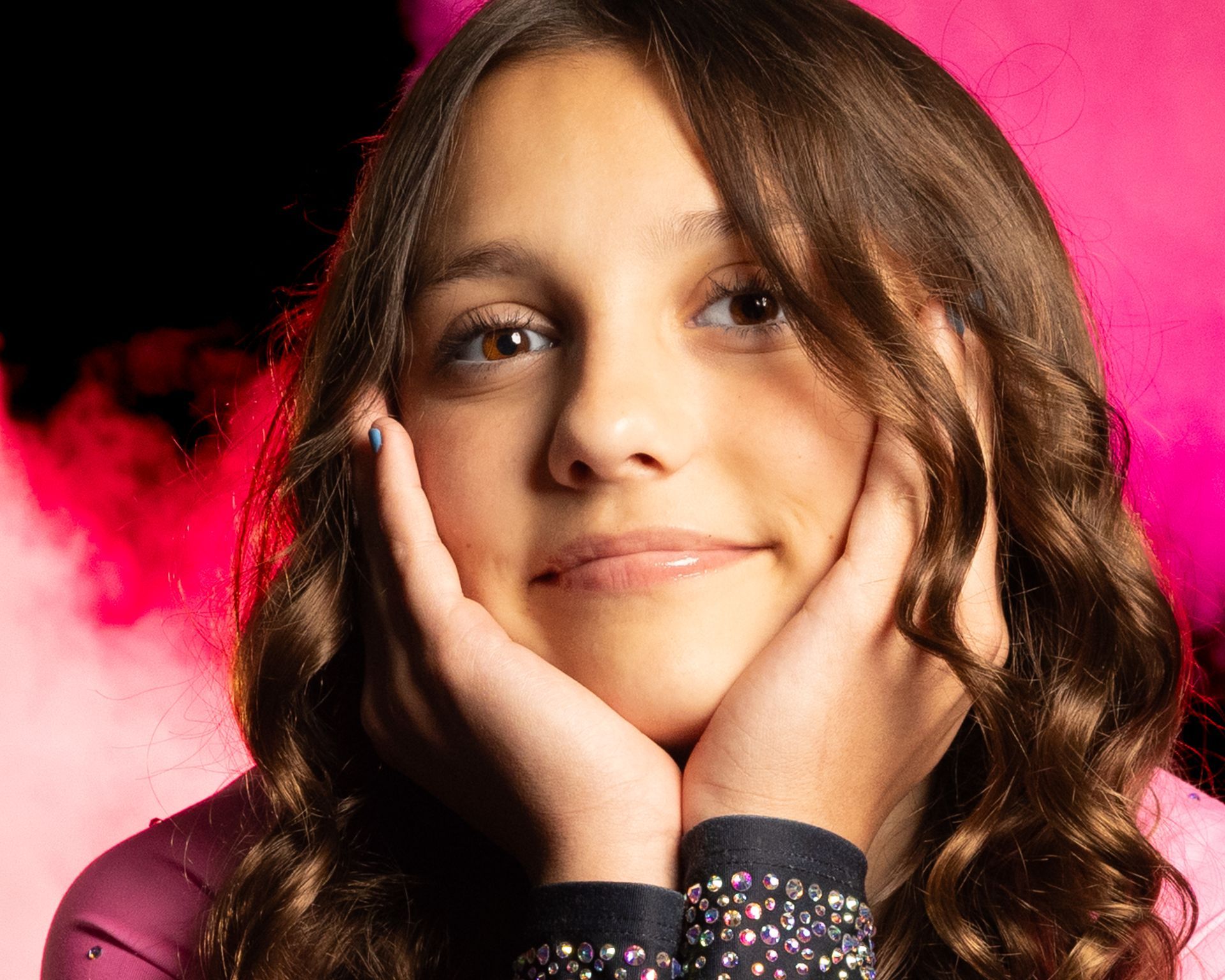 A girl with light brown hair smiles, hands framing her face; pink and red smoky background.