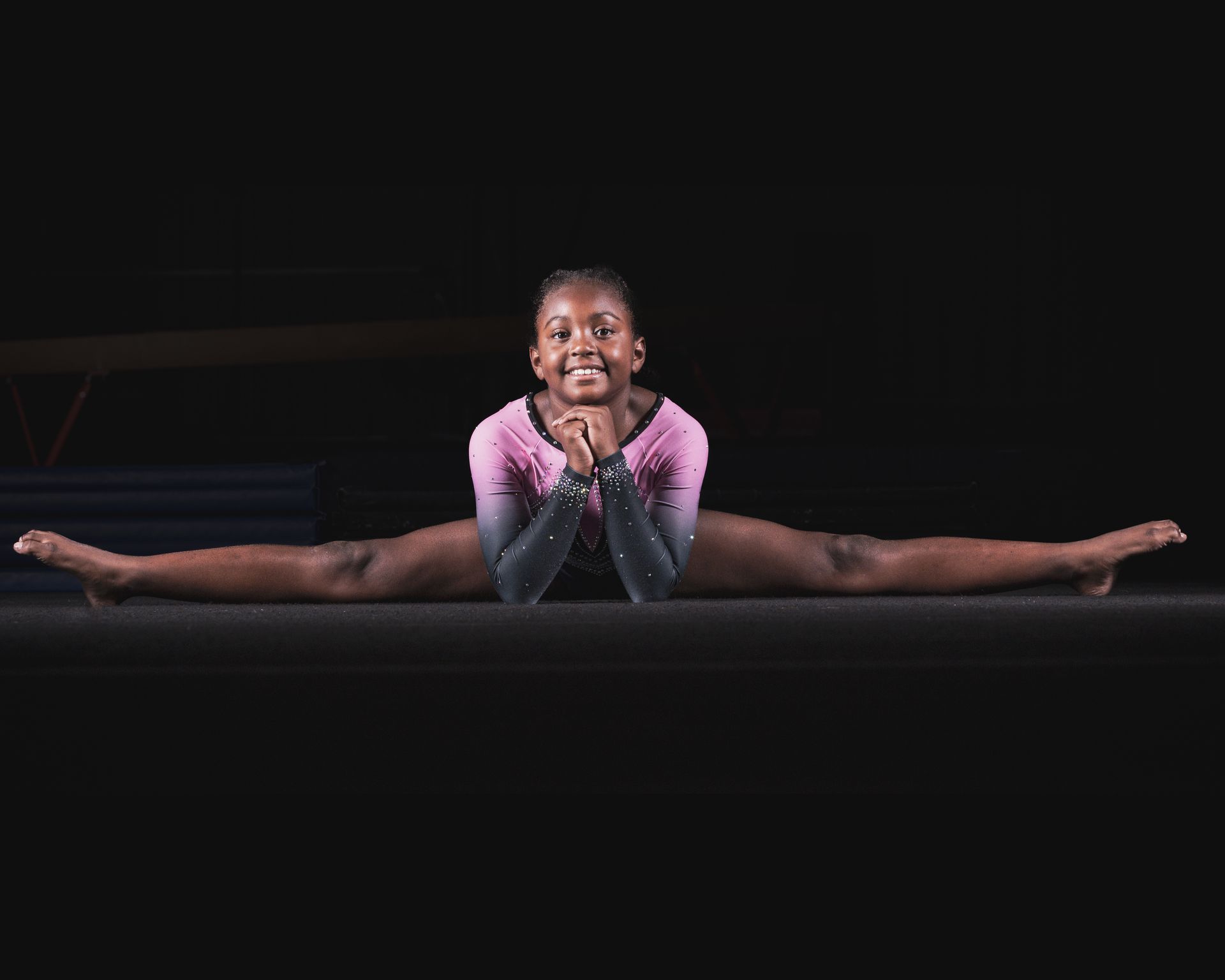 Gymnast in a full split, smiling, wearing a pink and black leotard, on a dark mat.