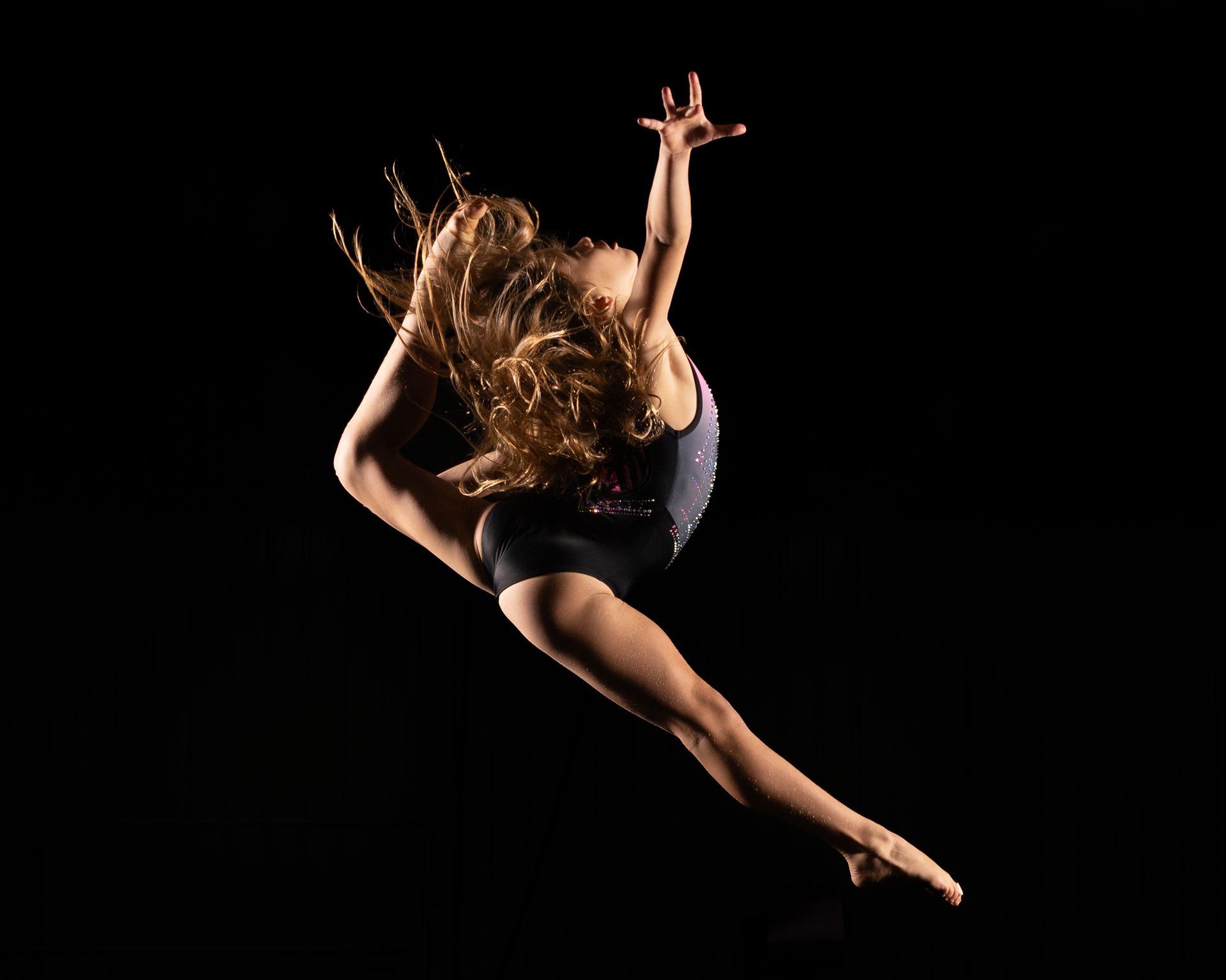 Dancer in black leotard leaps, arm extended, hair flowing, against black background.