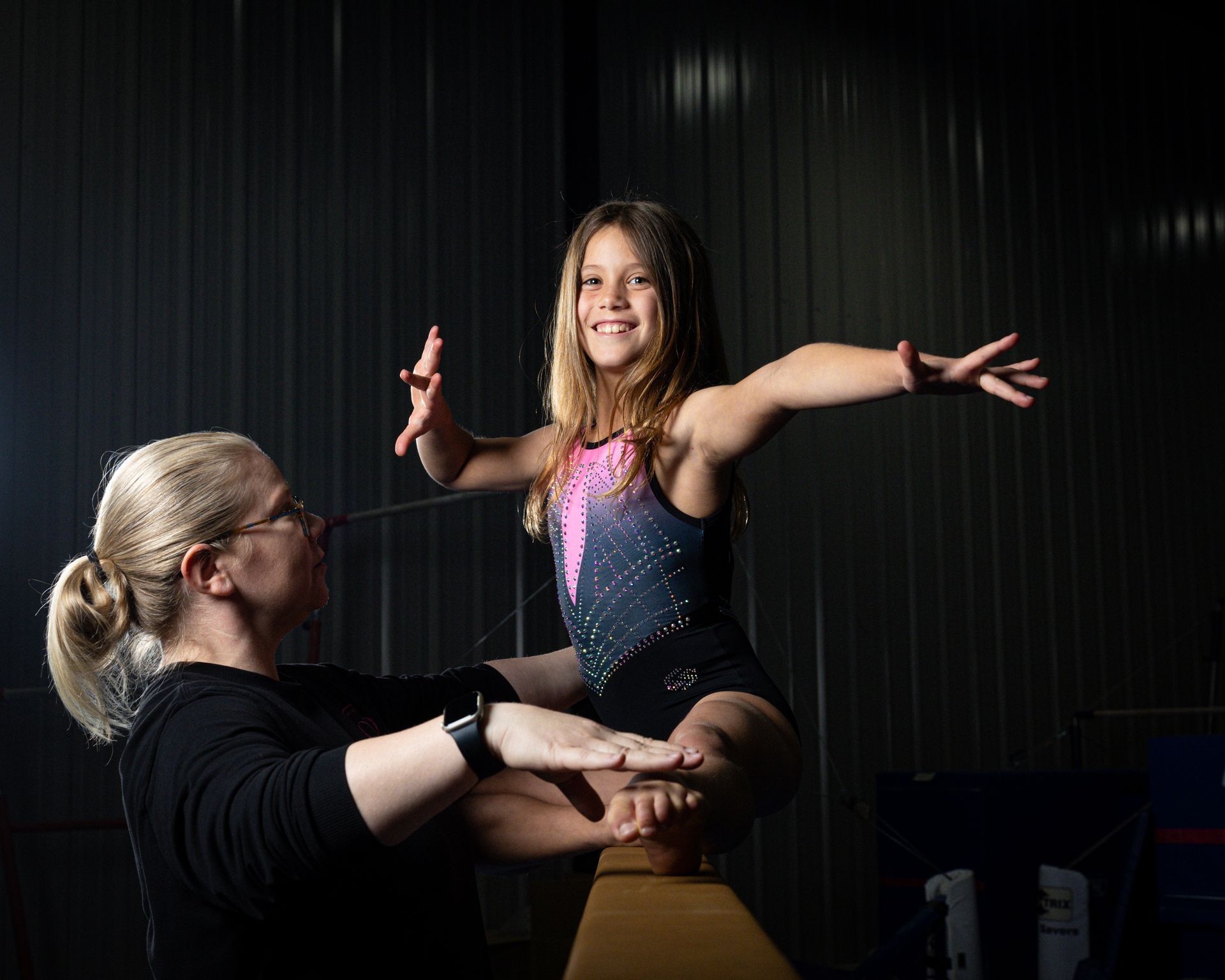 Girl on balance beam, arms outstretched, smiling. Coach assisting. Gym setting, dim lighting.