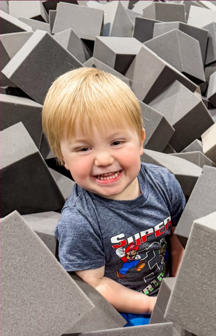Child with blonde hair smiling in a foam cube pit, wearing a superhero shirt.
