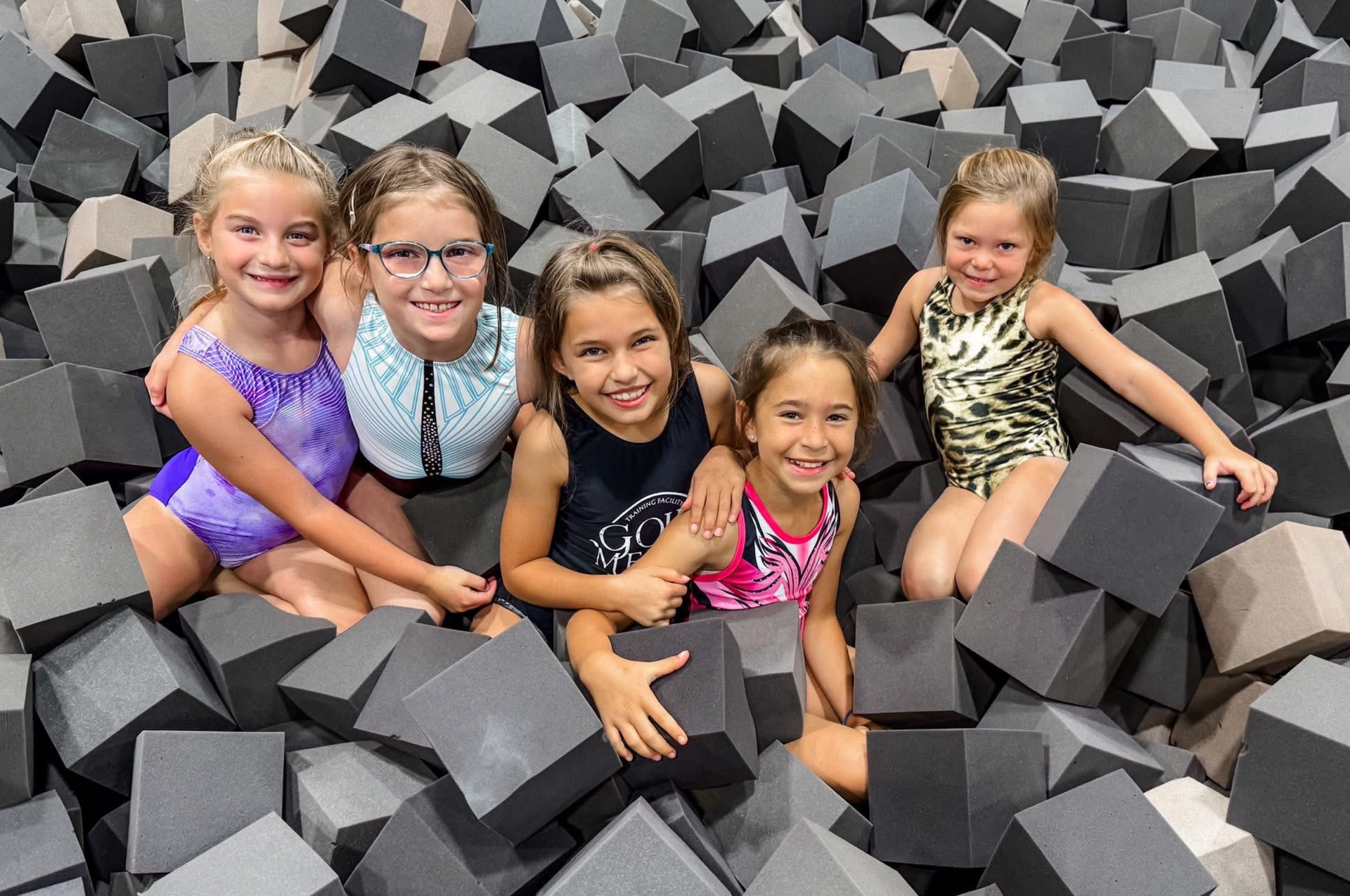 Five girls in gymnastics attire smiling, posing in a foam cube pit.