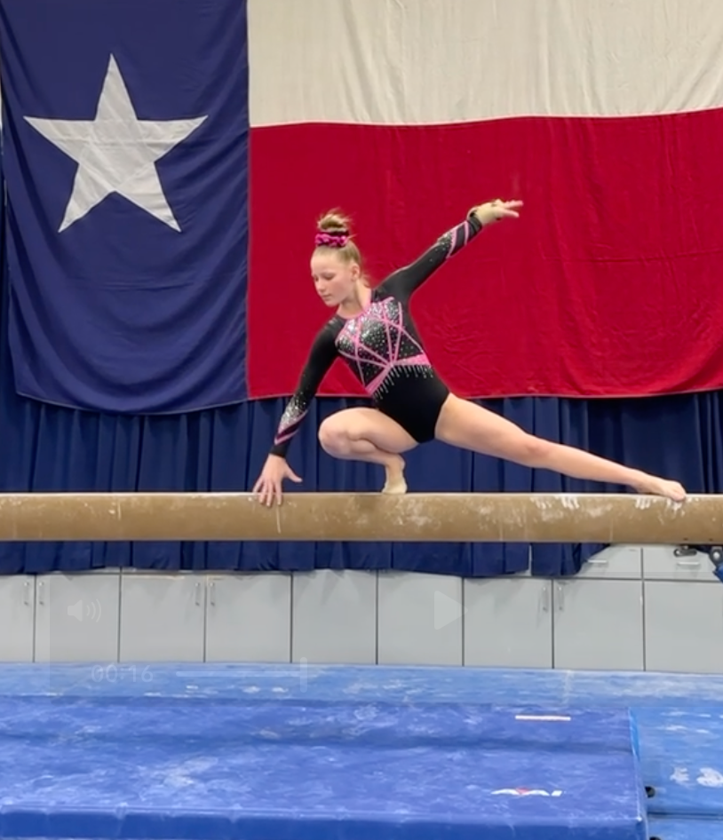 Gymnast balances on a beam, extending one leg. She wears a black and pink leotard in a gym with a Texas flag.