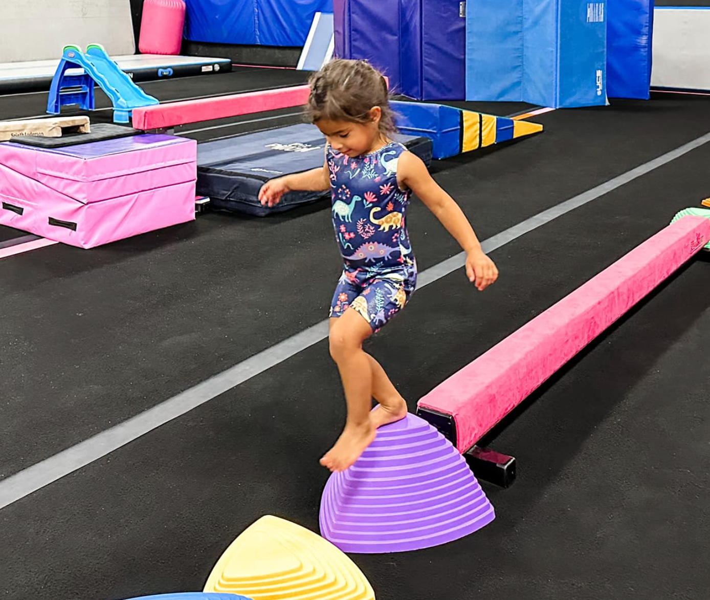 Child in gymnastics attire balances on purple stepping stones and a pink beam in a gym.
