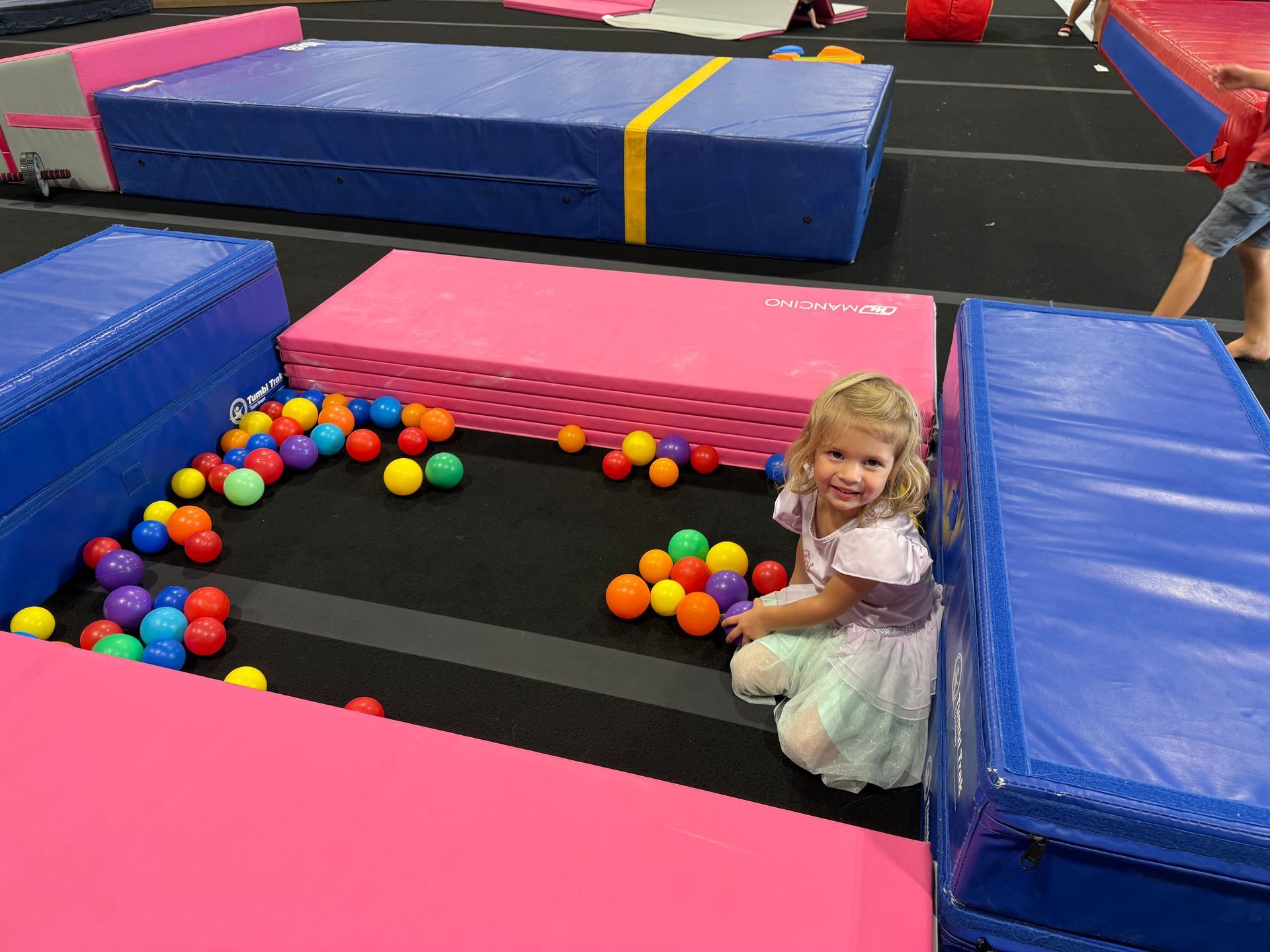 Young child in a princess dress sits near colorful balls and gym mats, smiling.