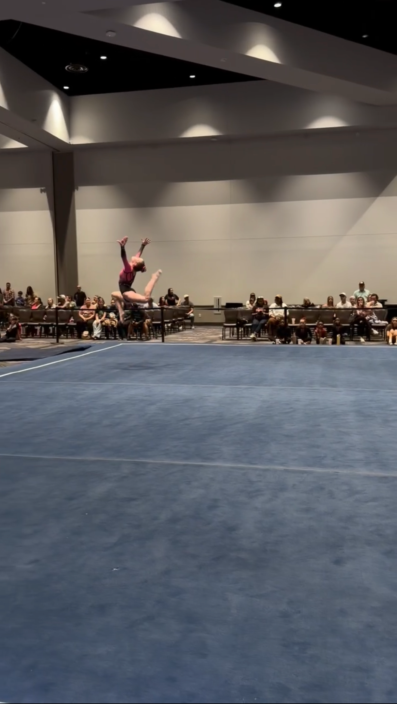 Gymnast performs a flip in the air on a blue mat, indoors in front of an audience.