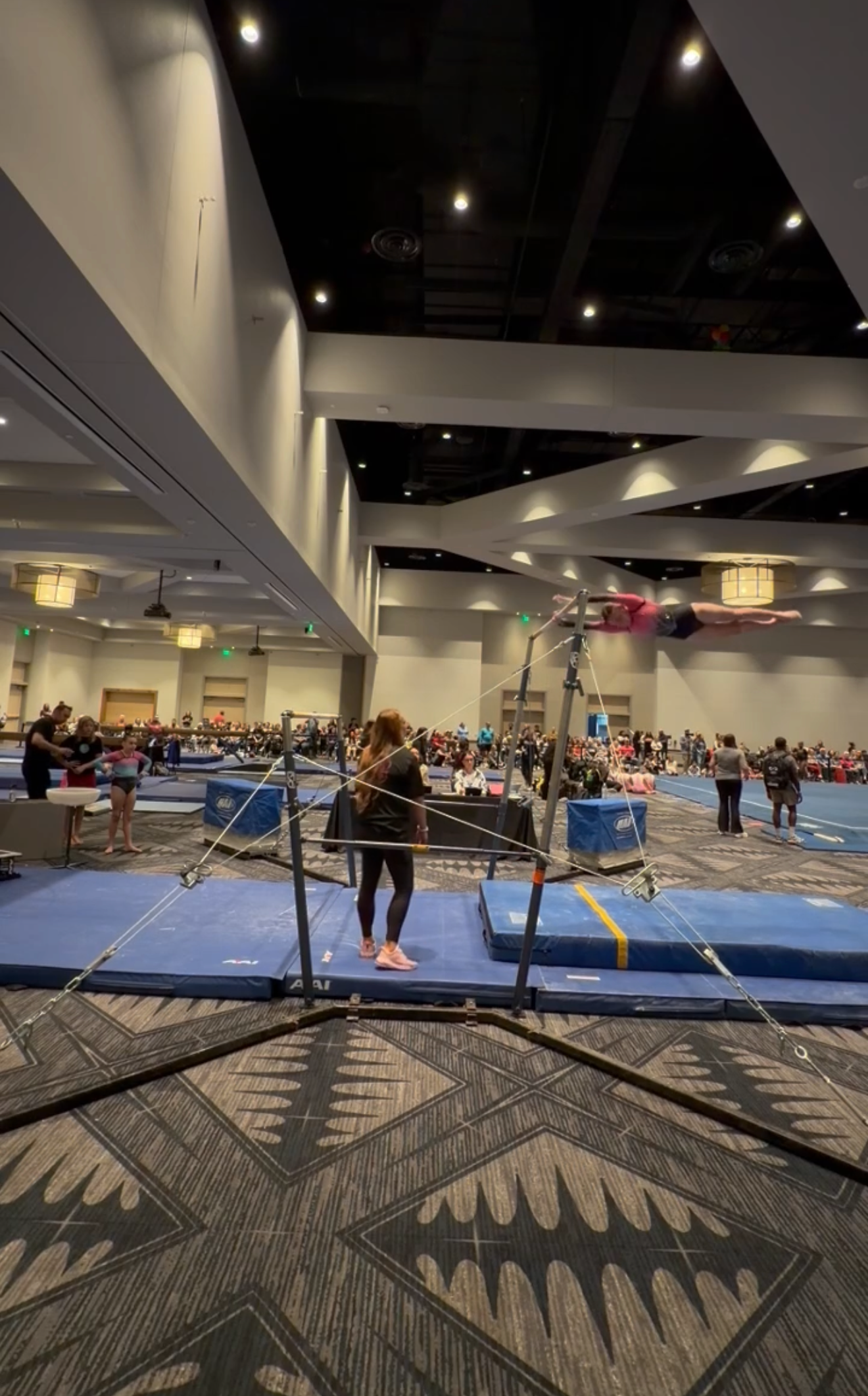 Gymnast stands near uneven bars, in a large gym. Other gymnasts are in the background, near other equipment.