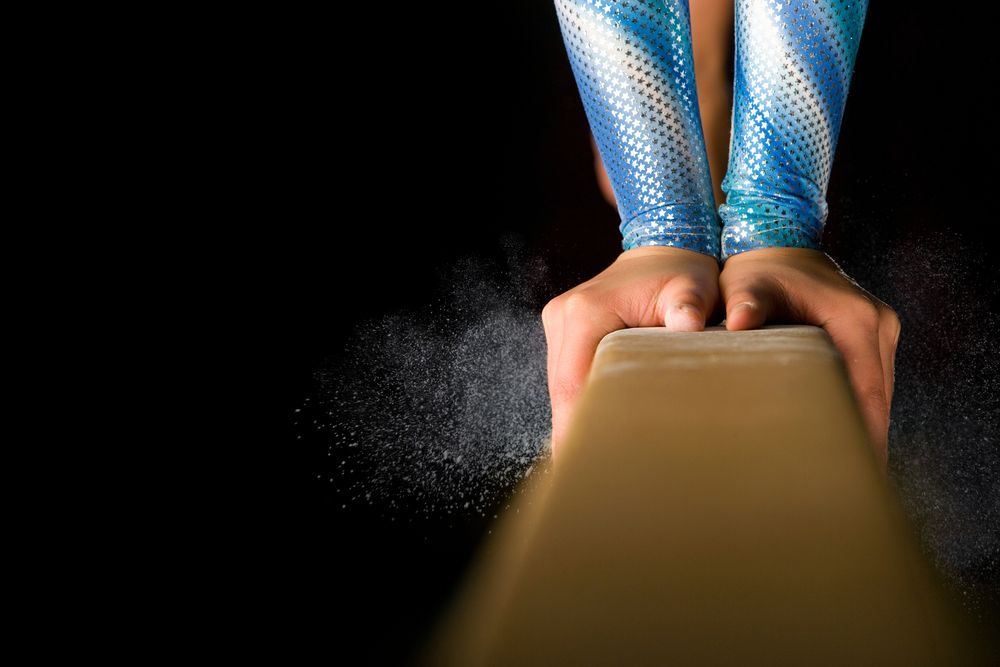 Gymnast's hands gripping a balance beam, chalk dust rising against a black background.
