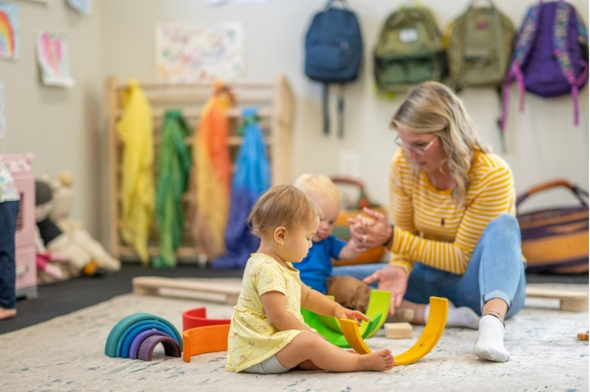 Child plays with toys as teacher watches in a brightly-lit classroom.