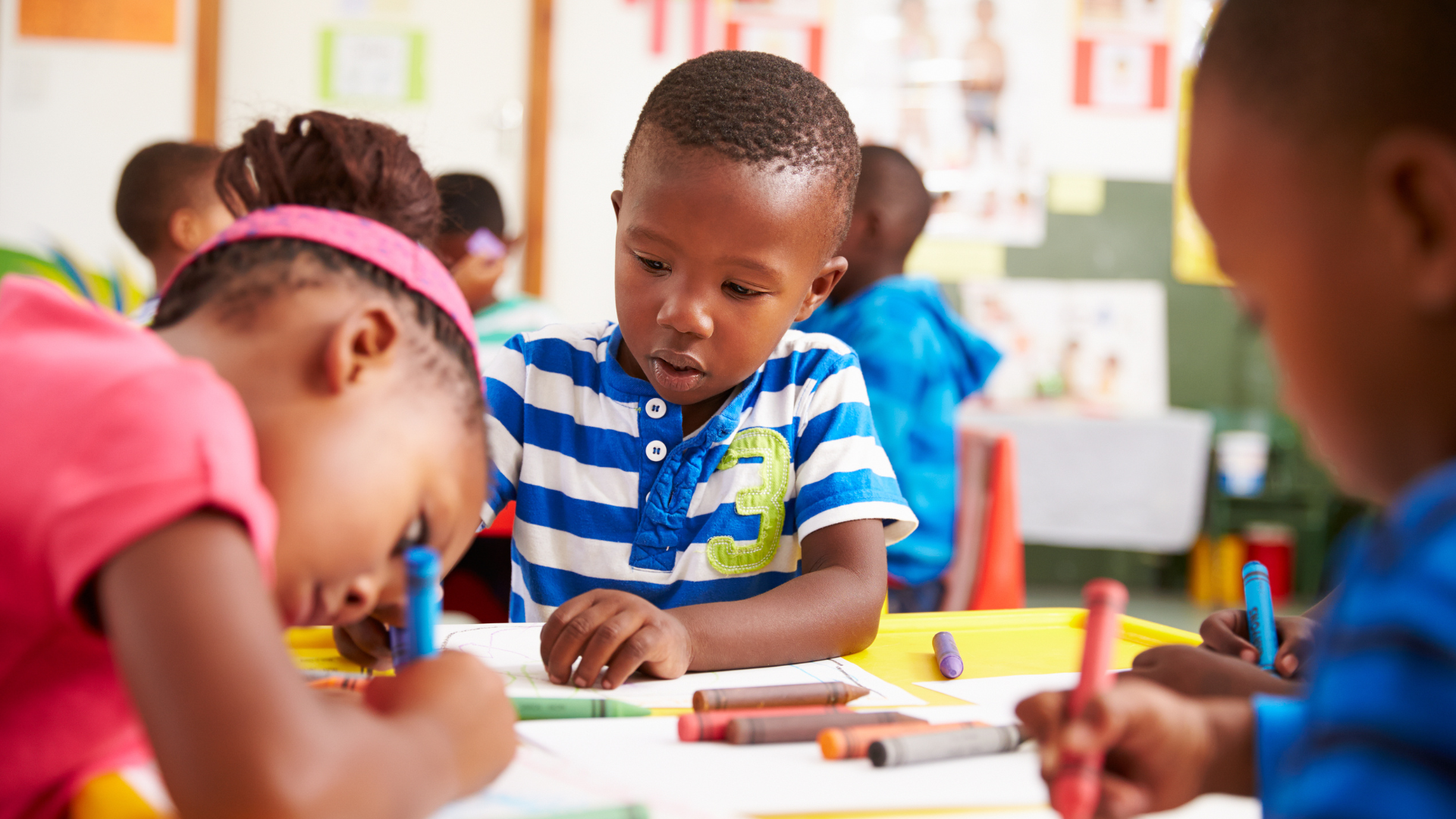 A group of children are sitting at a table in a classroom drawing with crayons.