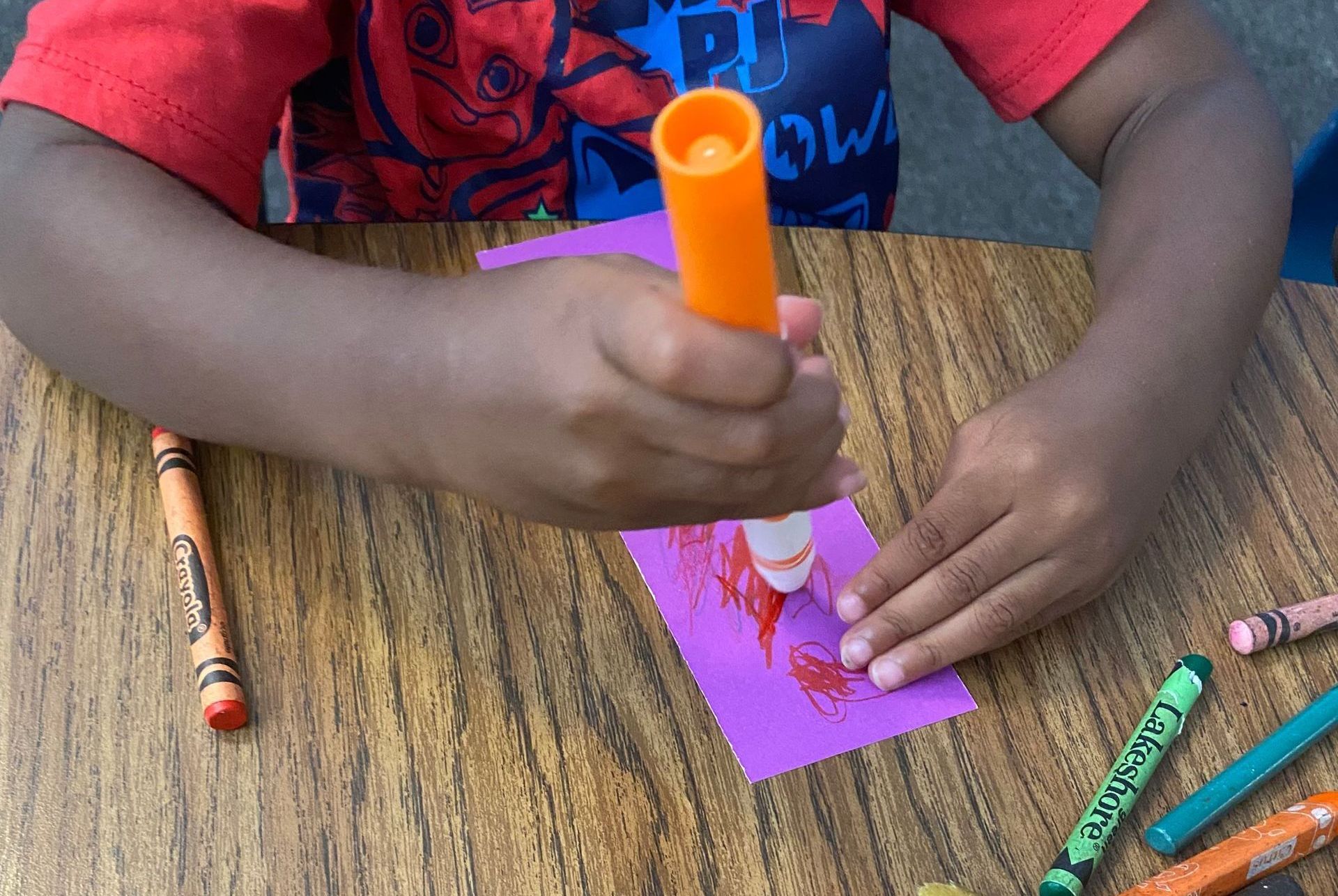 Child coloring with an orange marker on purple paper at a table, surrounded by crayons.