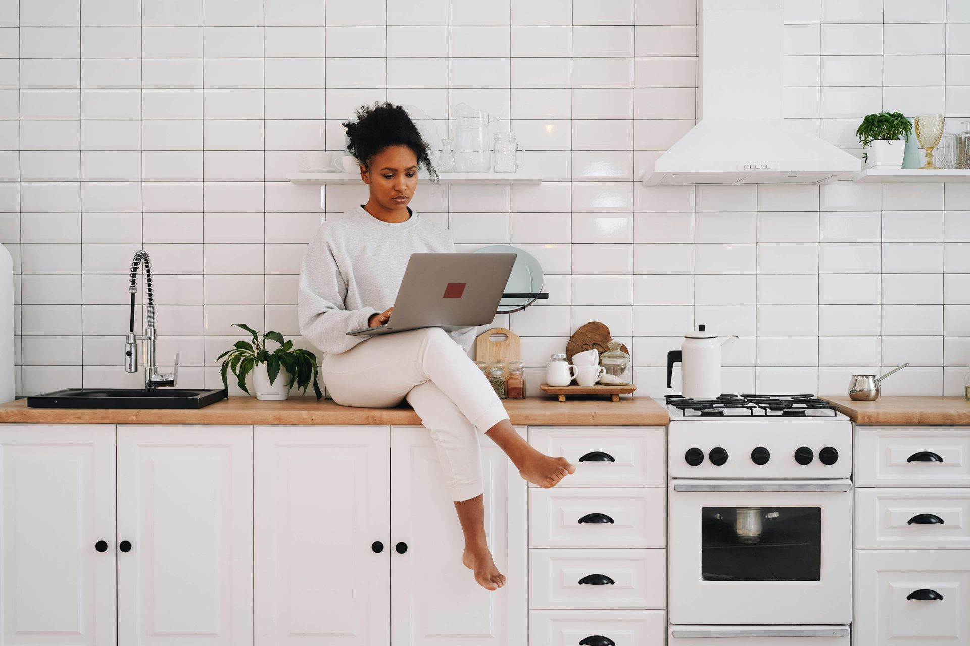 Een vrouw zit op het aanrecht in de keuken en gebruikt een laptop.