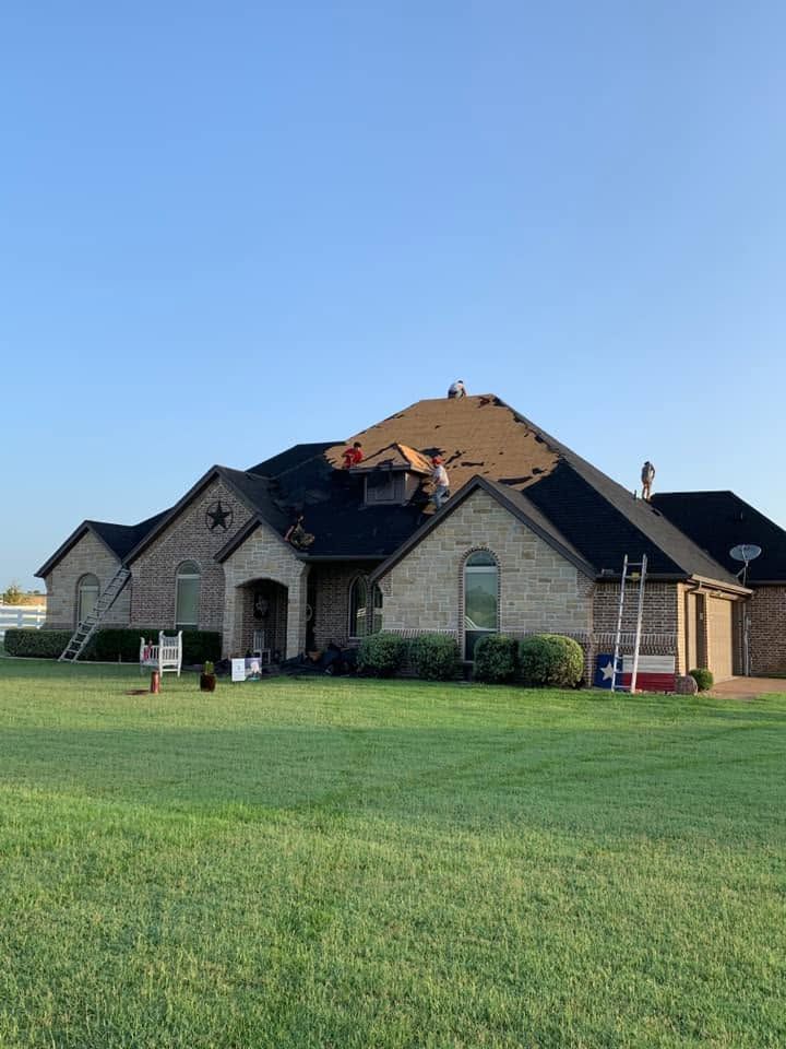 Damaged suburban house with torn roof shingles and debris on a green lawn under a clear blue sky