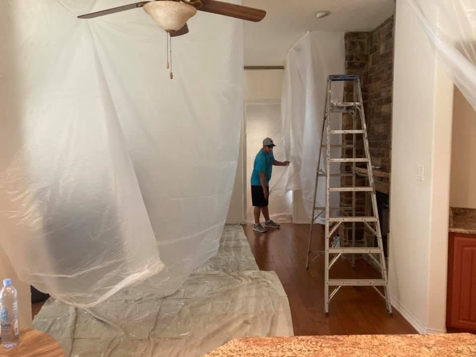 Person painting a room with plastic-covered furniture, ladder, and ceiling fan during home renovation
