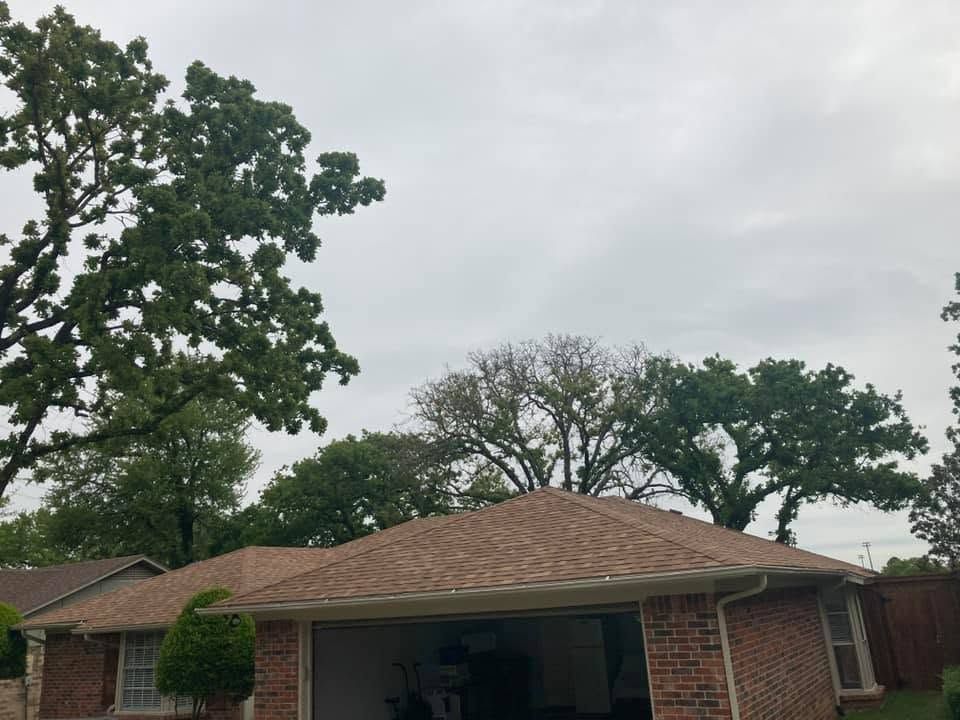 Brick house with brown shingle roof and large trees under a cloudy sky
