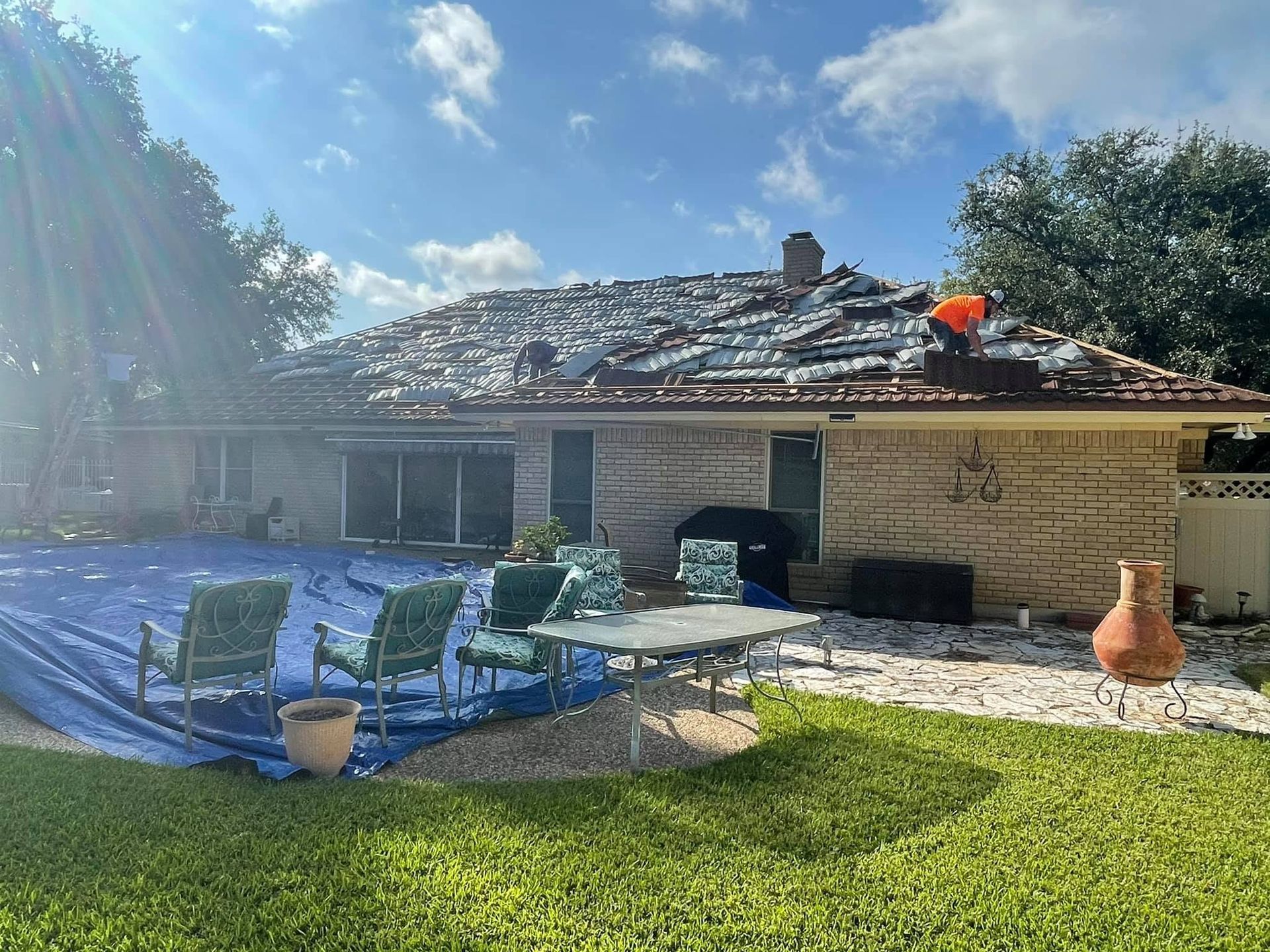 Single-story house with severe roof damage, blue outdoor chairs, and a sunny backyard after a storm