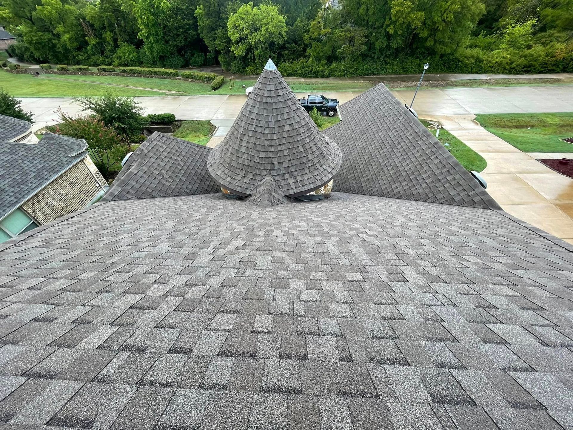 Gray shingled rooftops with a conical turret, viewed from above amid green trees and a road