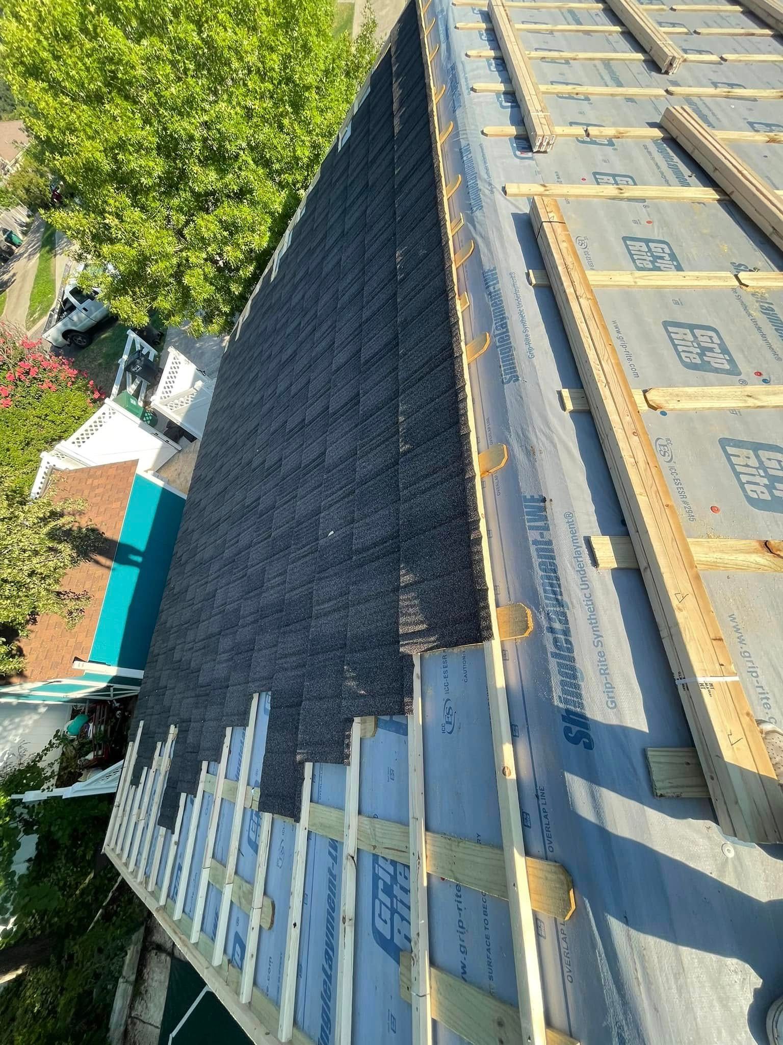 Roof under construction with dark shingles, wooden framing, and trees beside the building.