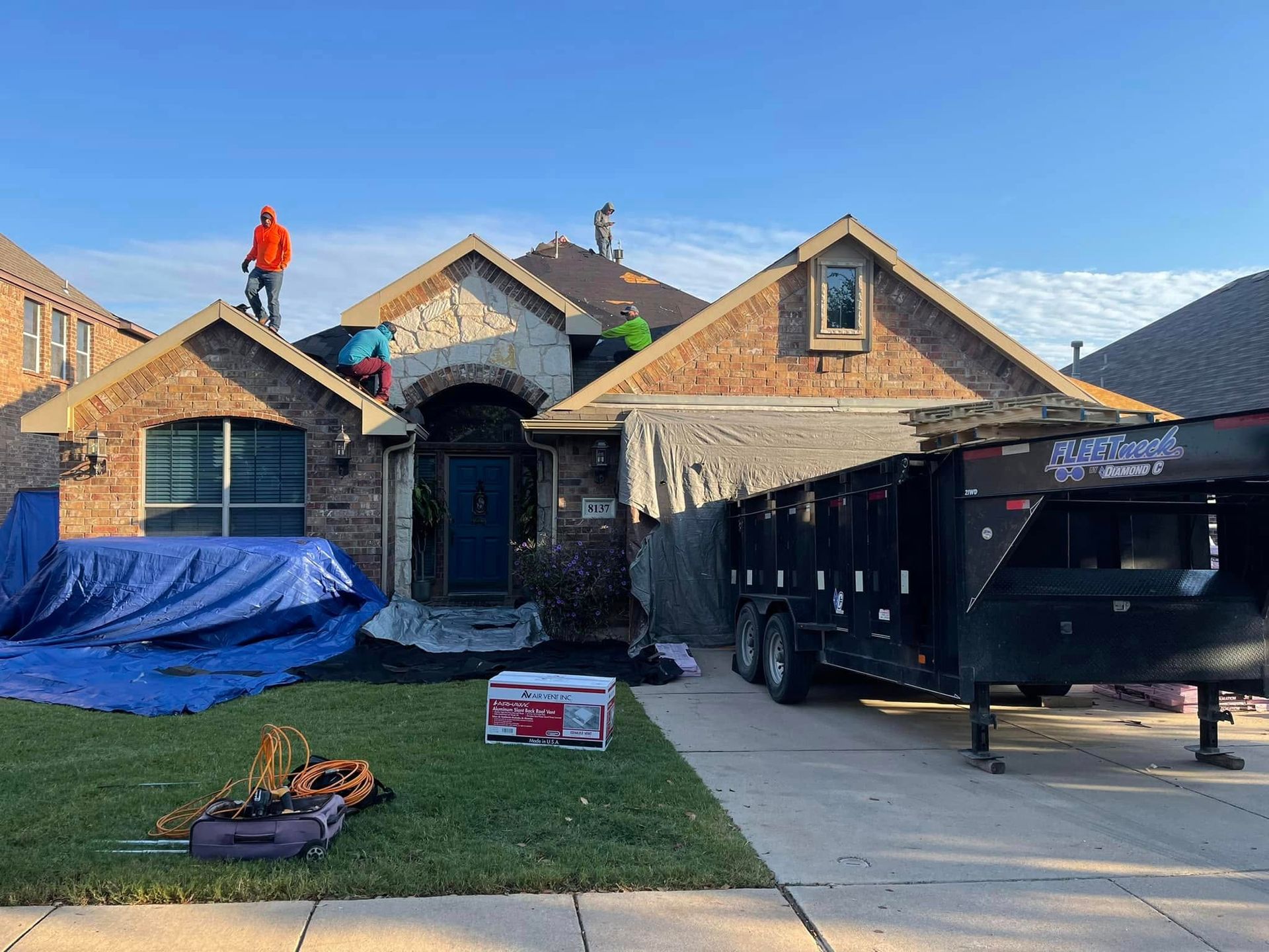 Workers roofing a brick house with blue tarp and a black dumpster in the driveway under a clear sky