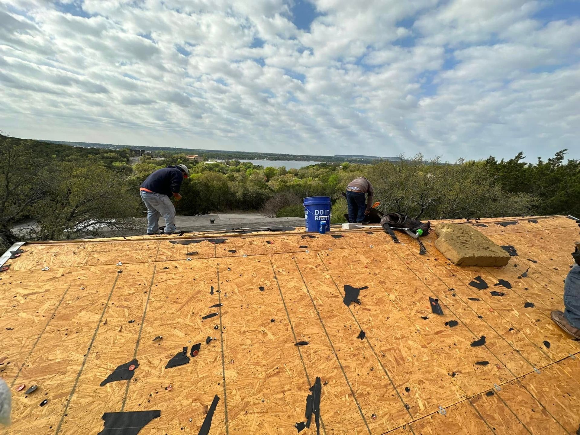 Roof under construction with two workers on plywood, tools, and blue bucket overlooking a wooded landscape