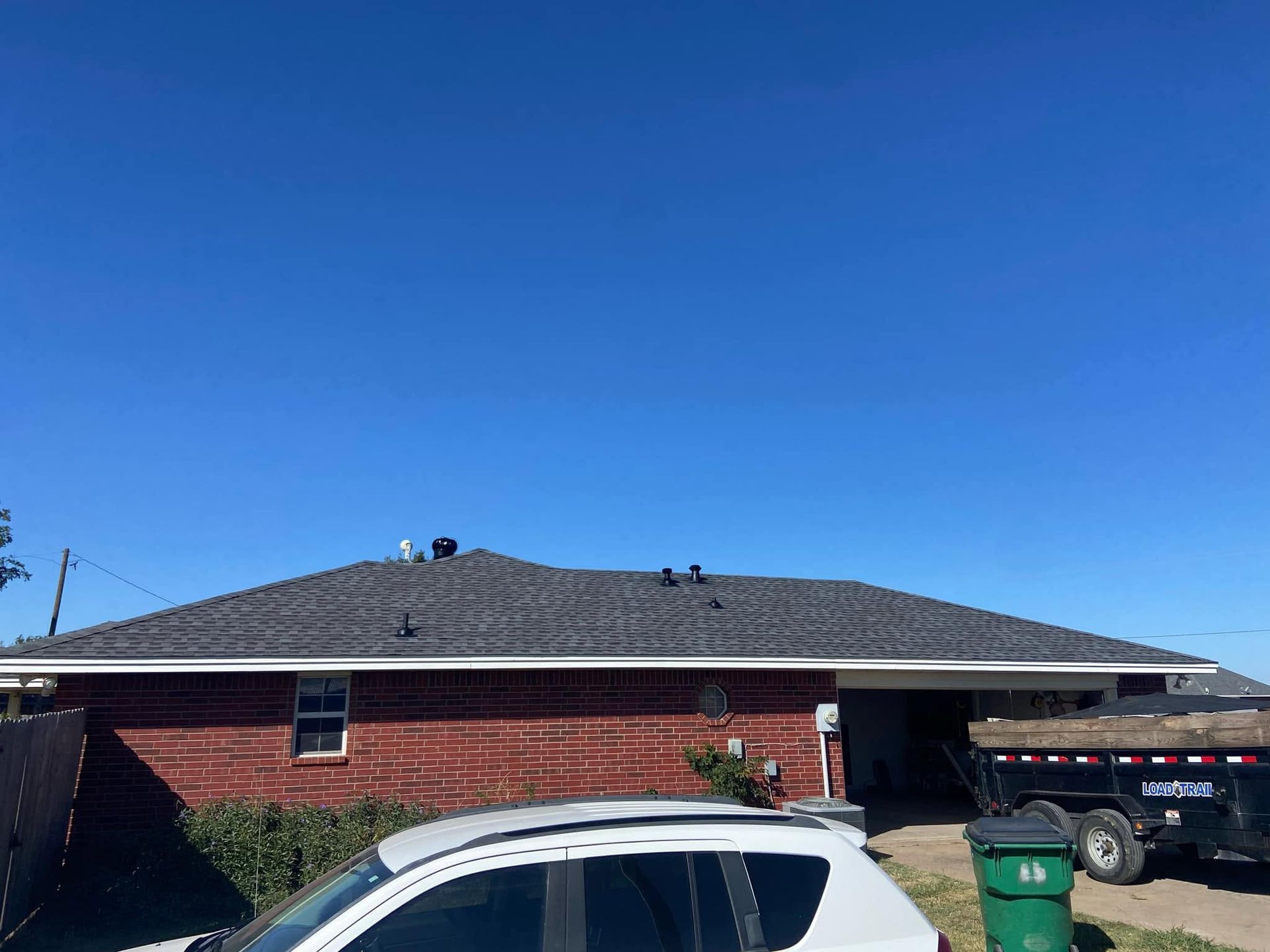 Brick house under a clear blue sky with a white car and green trash bin in the driveway
