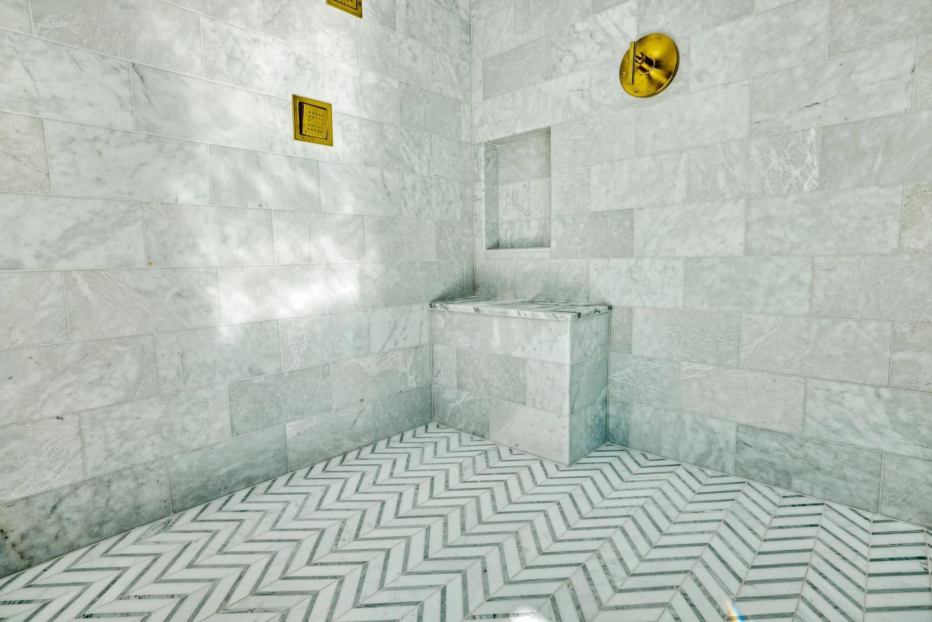 White tiled shower room with zigzag floor and gold fixtures, featuring a raised corner bench and shelf.