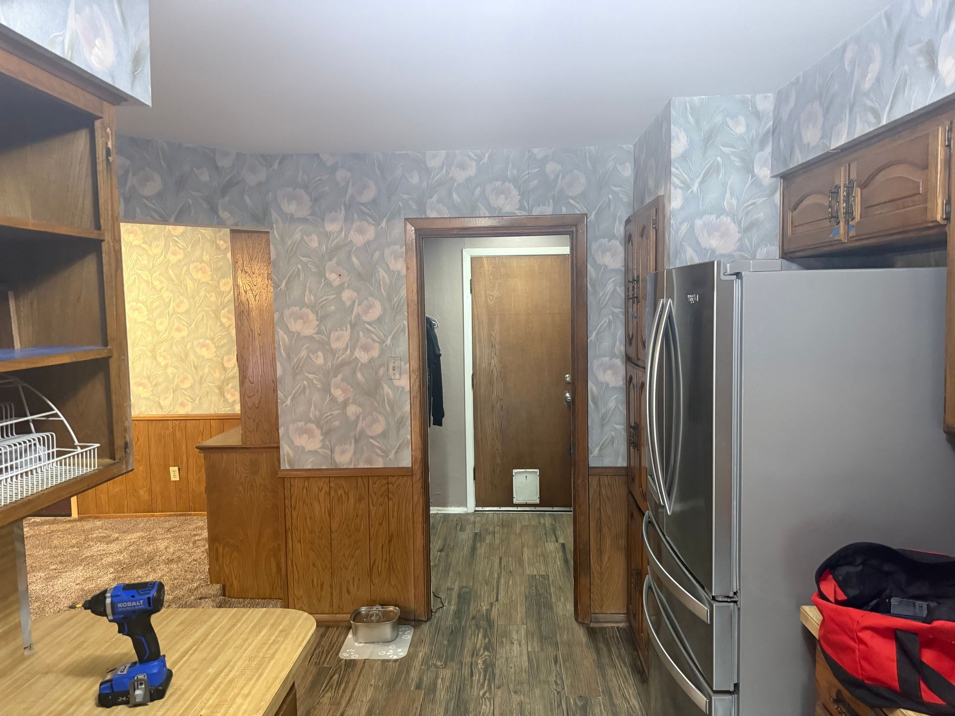 Small kitchen with wood cabinets, floral wallpaper, stainless fridge, and a blue drill on the countertop.