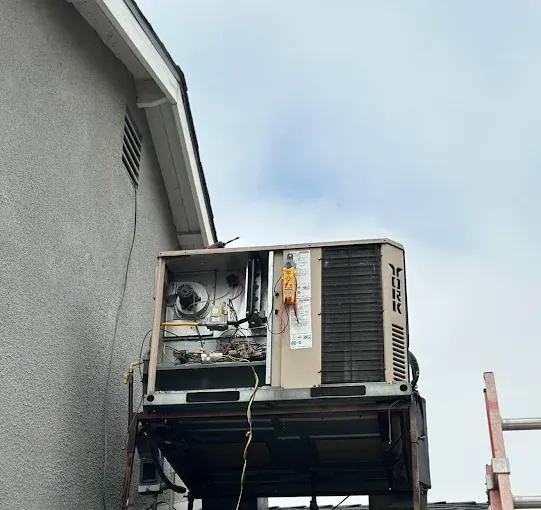 Damaged rooftop HVAC unit with exposed wiring on a building exterior, against a cloudy sky