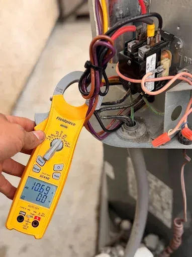 Hand holding yellow digital multimeter testing wiring inside an electrical unit with exposed components