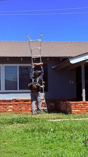 Person standing on a ladder under another person being lifted outside a house, with green lawn and blue sky