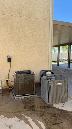 Two outdoor HVAC units beside a beige wall, with water puddled on the concrete.