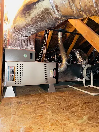 Attic HVAC unit with ductwork and insulated pipes on plywood flooring