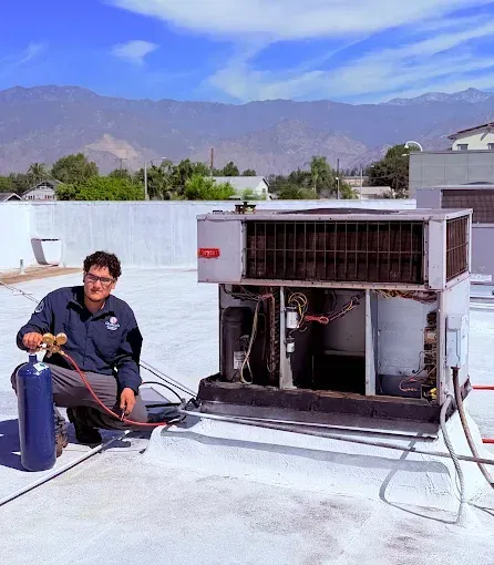 Technician kneeling on a rooftop beside HVAC equipment, holding tools and a gas cylinder with mountains in the background