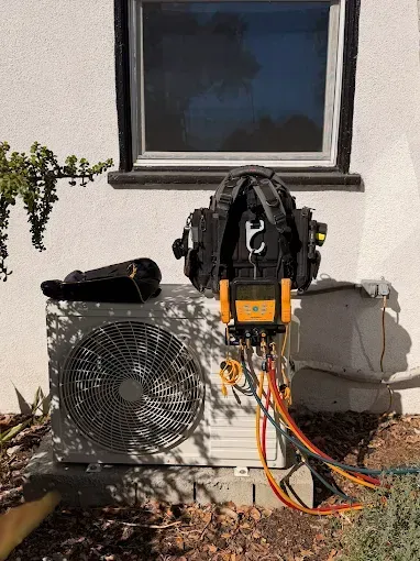 Outdoor HVAC unit with exposed wiring and a wall-mounted electrical device beneath a window