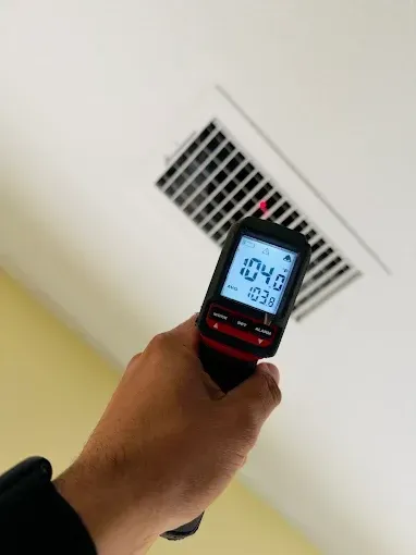 Hand holding a digital thermometer near a ceiling vent, showing temperature readings.